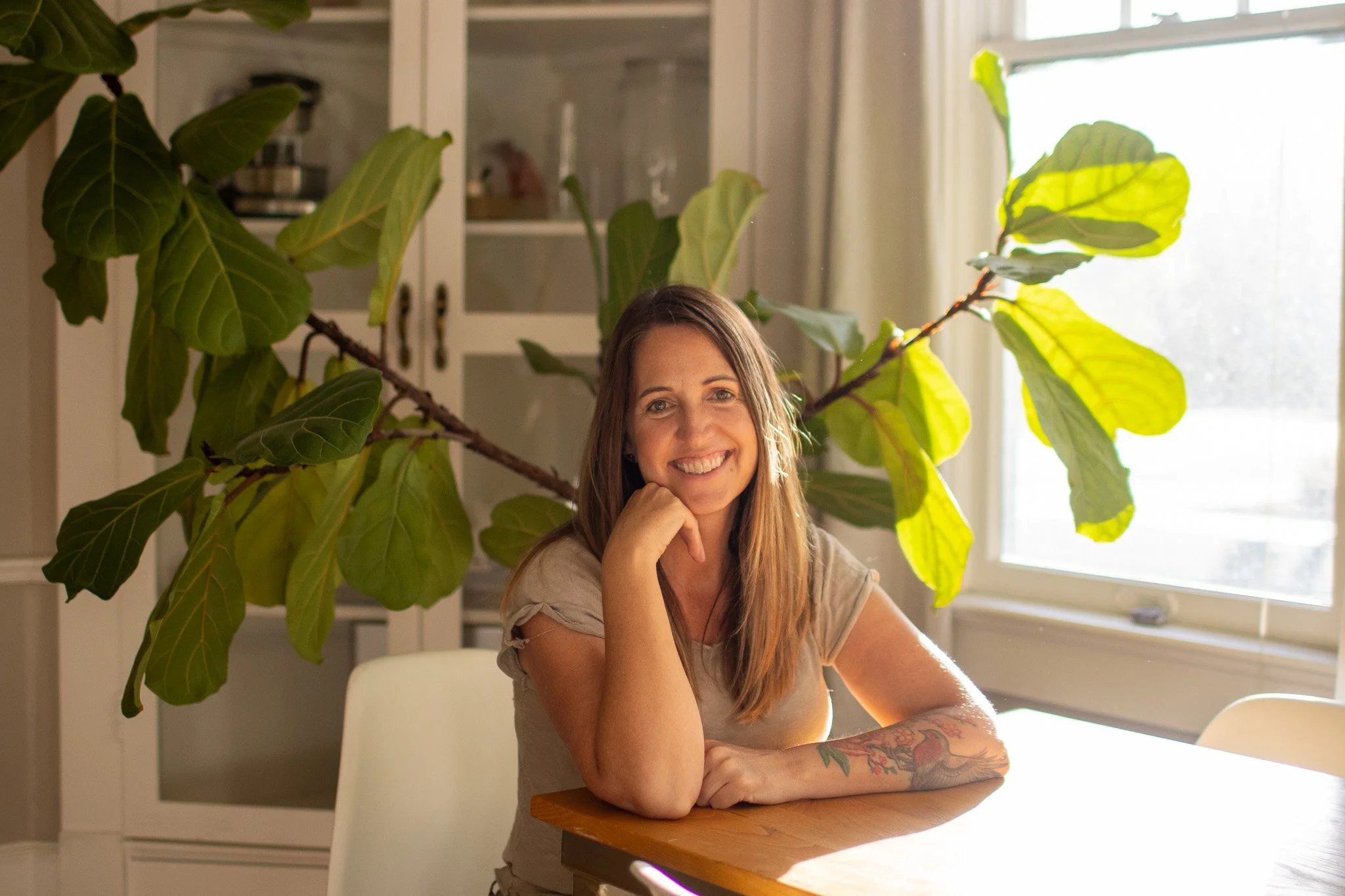 A woman with long brown hair and a tattoo on her left arm smiling at the camera, sitting at a wooden table in a well-lit room with a large plant and a window behind her.