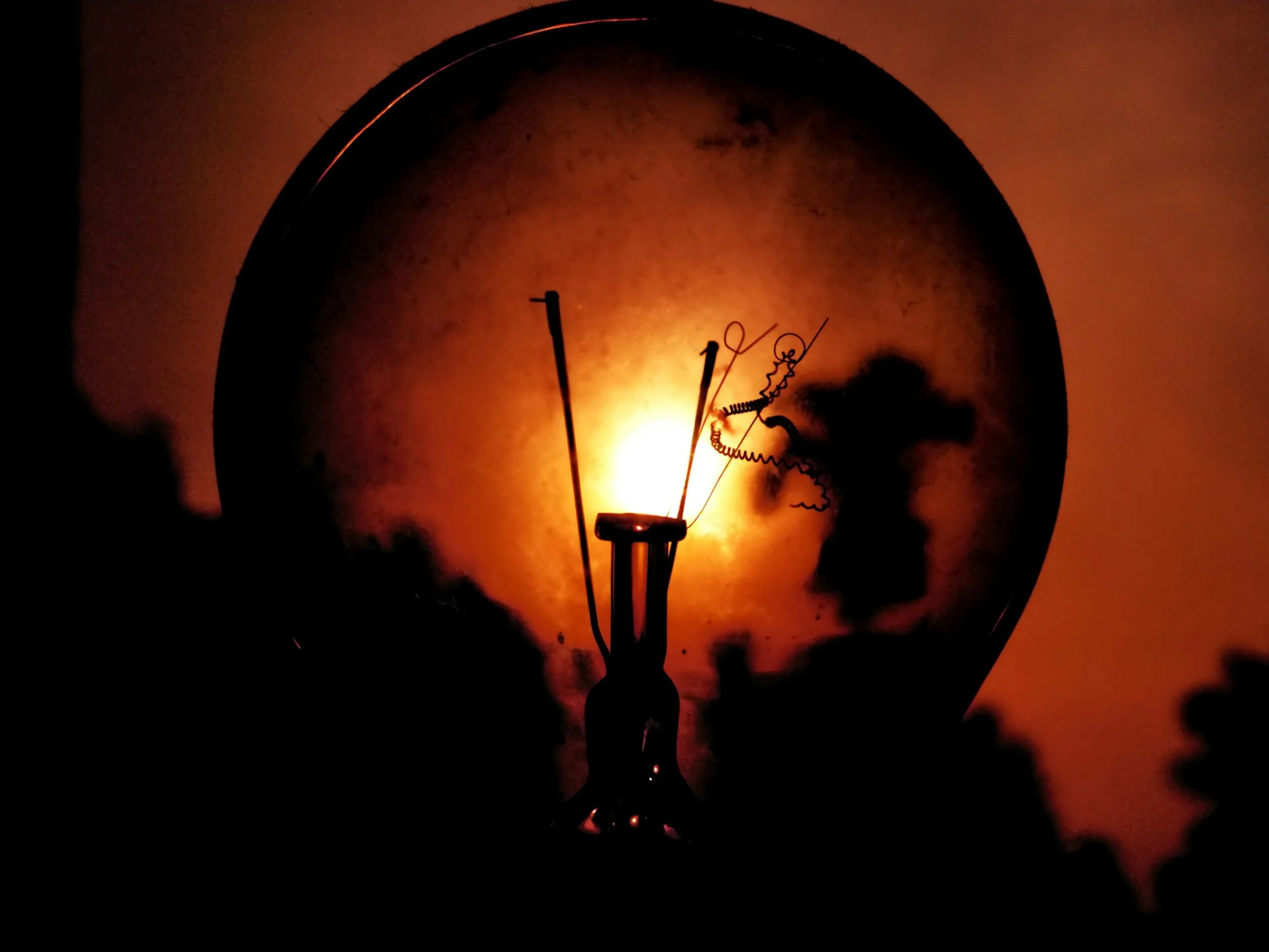 Close-up of an incandescent light bulb filament lit up, casting a shadow on the glass surface, with a warm orange glow.