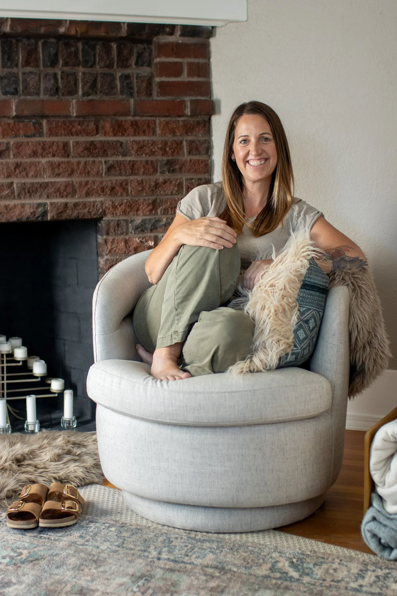 A woman sitting cross-legged on a light-colored, round armchair next to a brick fireplace, smiling and holding a fluffy dog. There are shoes, candles, and a rug visible in the room.