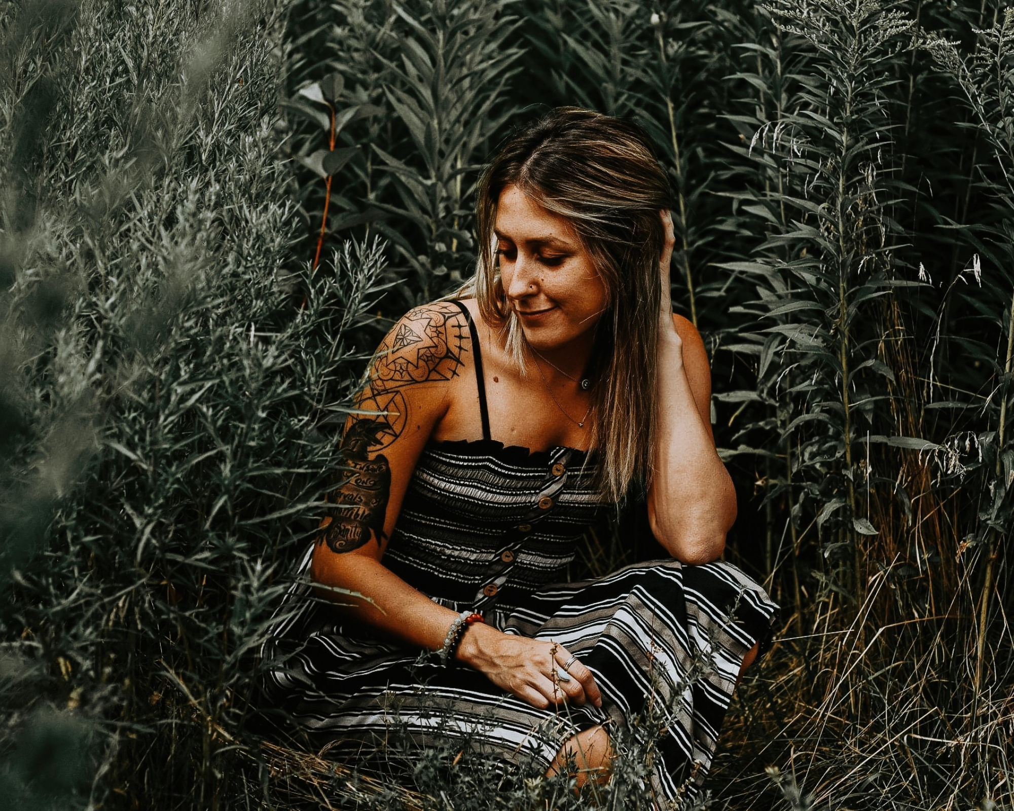 Person sitting in tall green plants, looking down with a relaxed expression, wearing a striped dress.