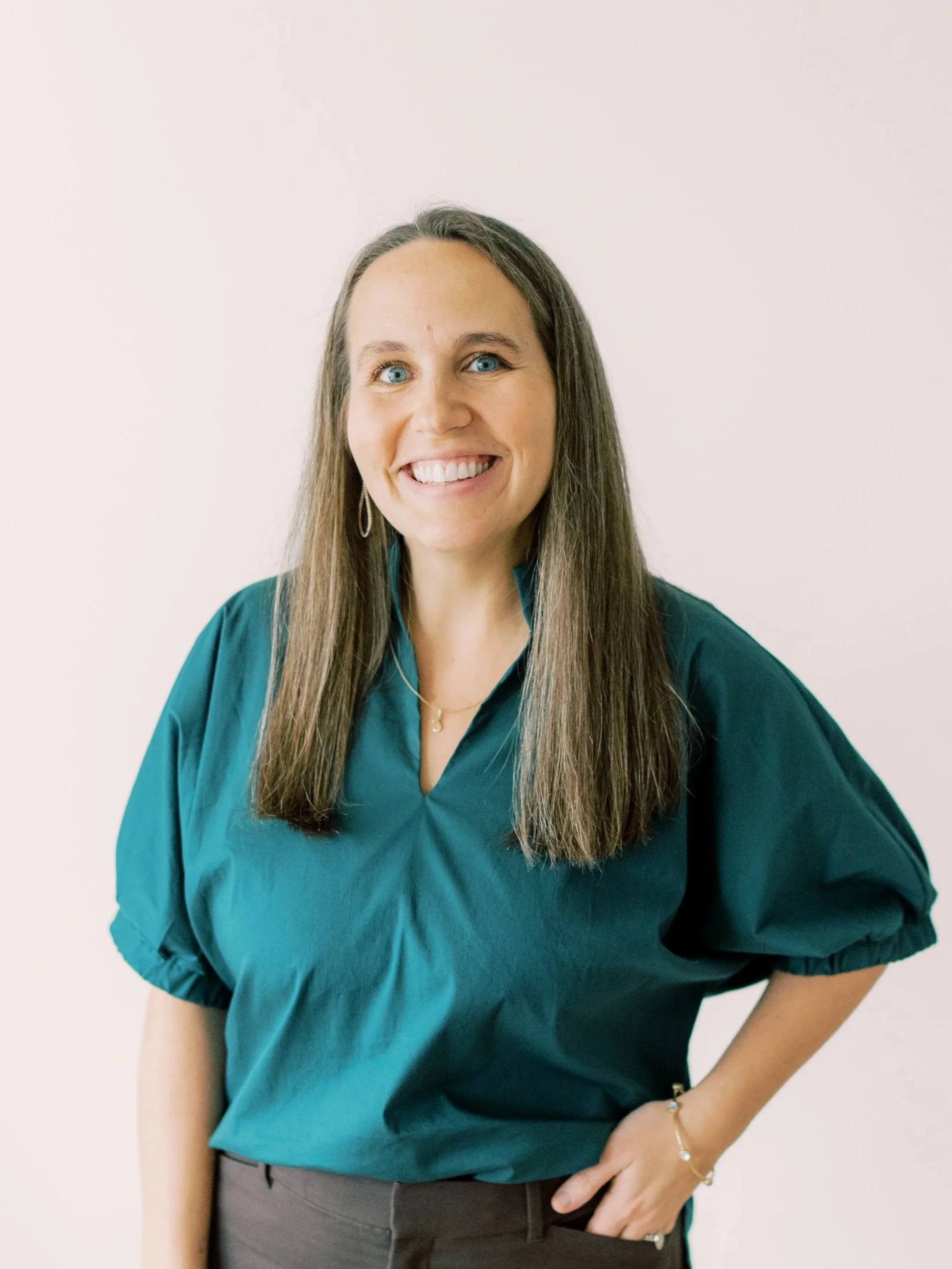 Smiling woman with long brown hair, blue eyes, wearing a teal blouse and jewelry, standing against a plain white background.