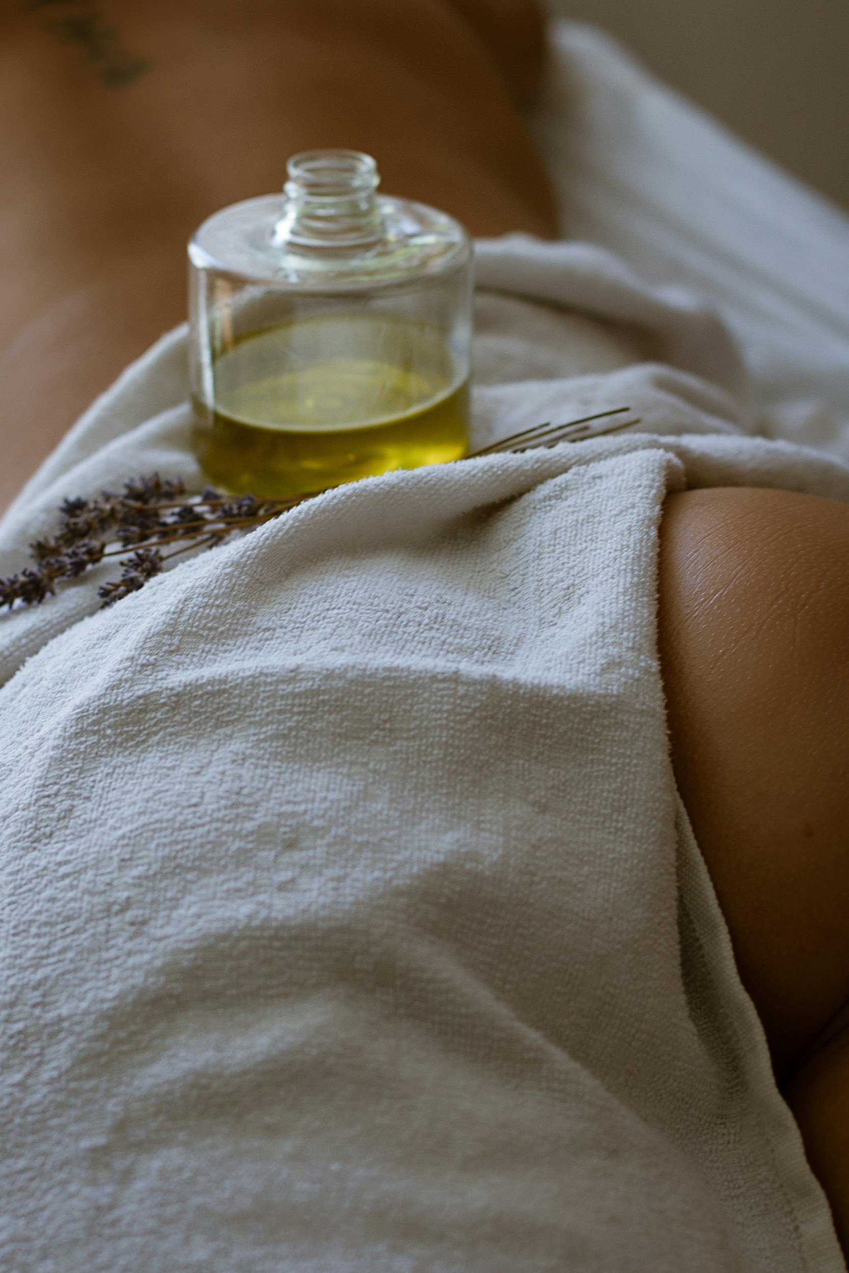 A close-up of a person's abdomen with a small glass bottle of massage oil resting on white towel, along with a sprig of lavender.