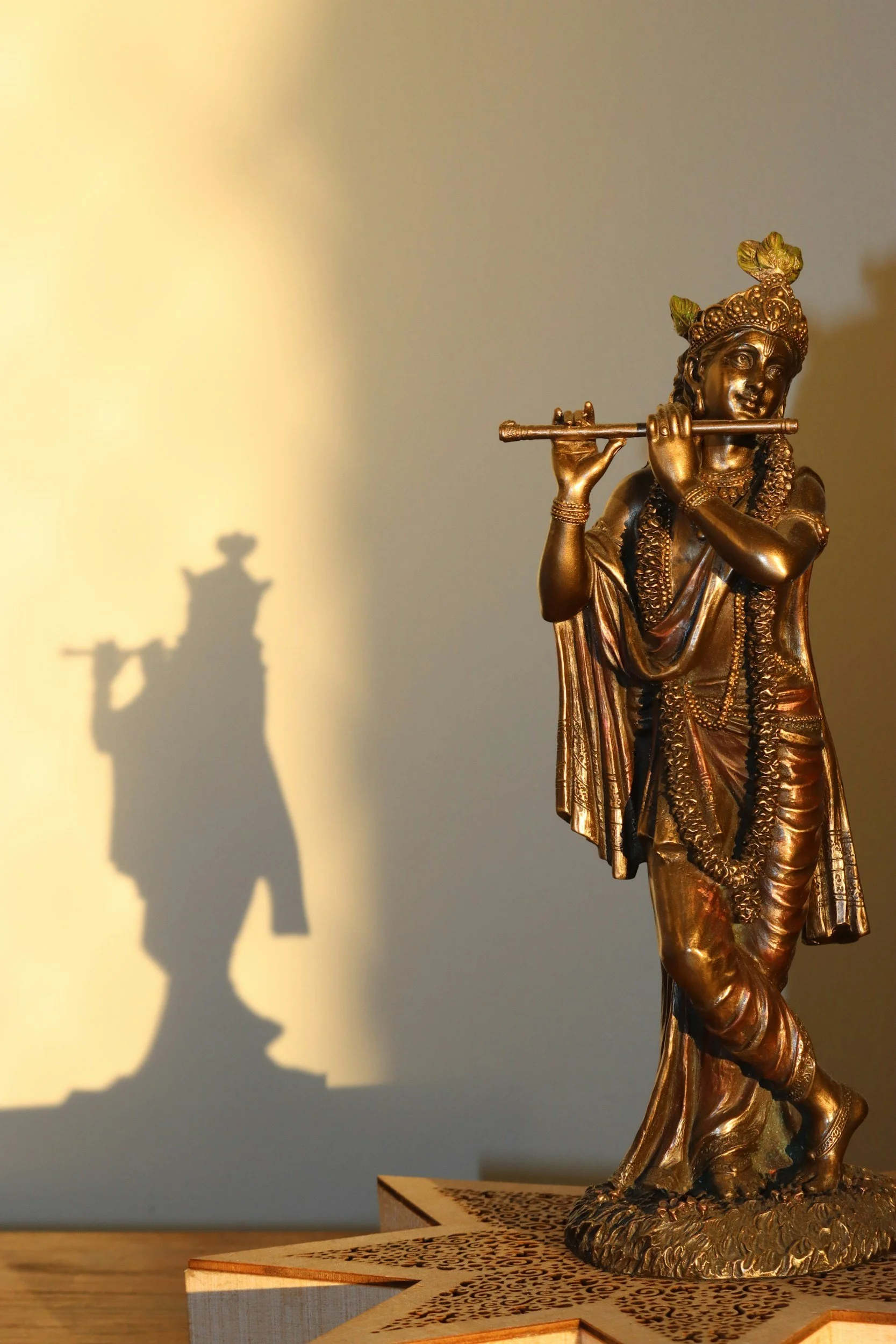A bronze statue of Lord Krishna playing flute, with a shadow of the statue cast on the wall behind it.