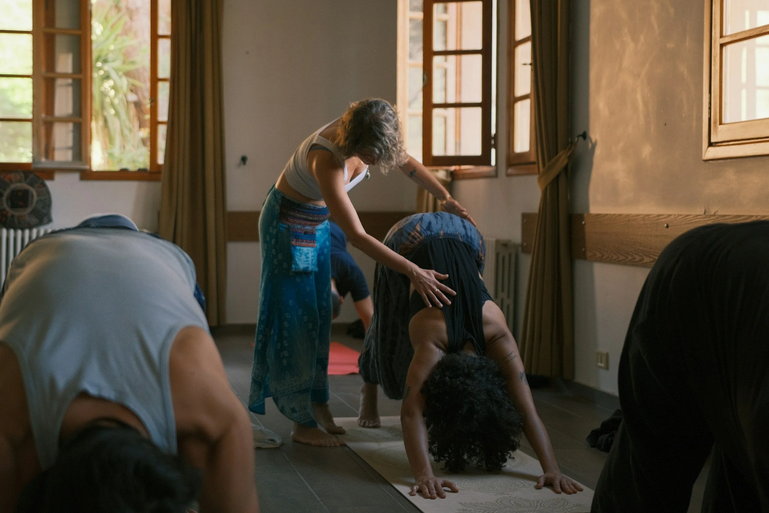 A woman leading a yoga class, guiding students through a pose in a sunlit room with large open windows.