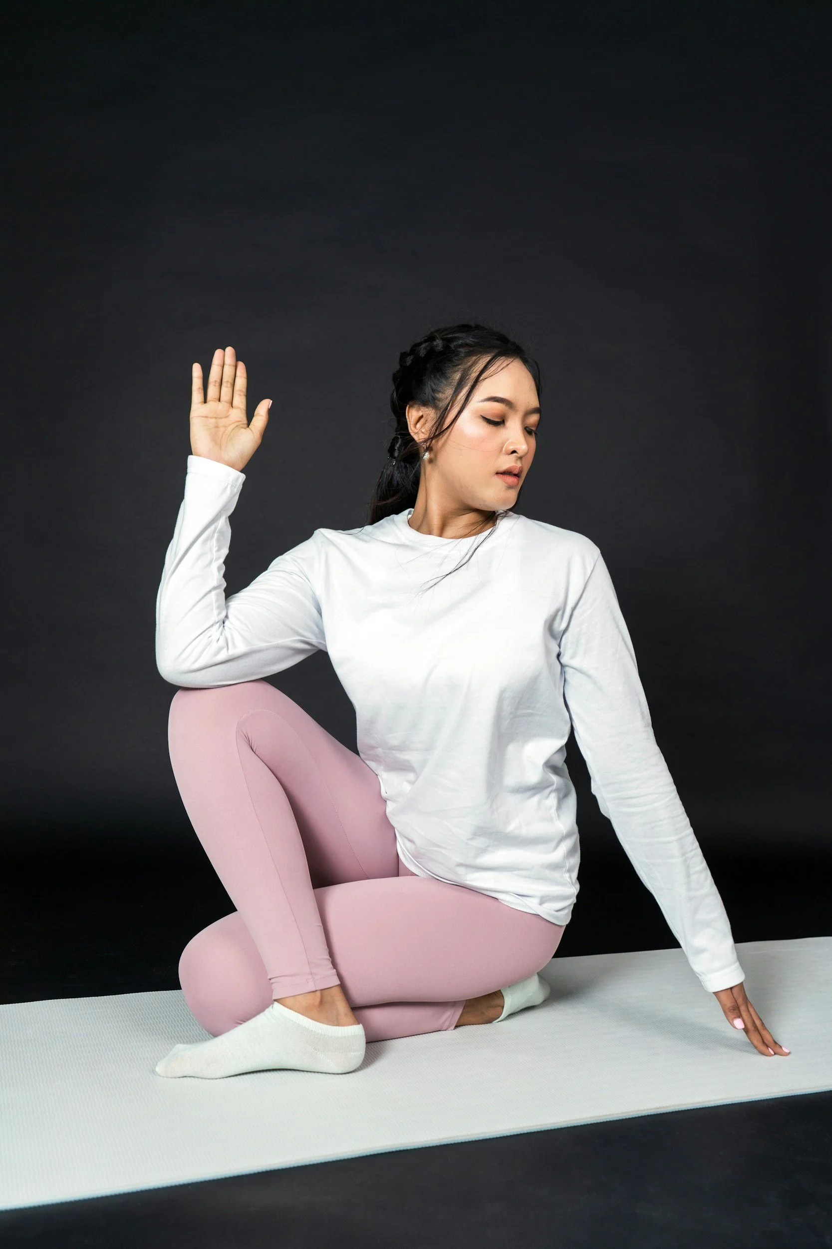 Woman in white long sleeve shirt and pink leggings practicing yoga on white mat against black background.