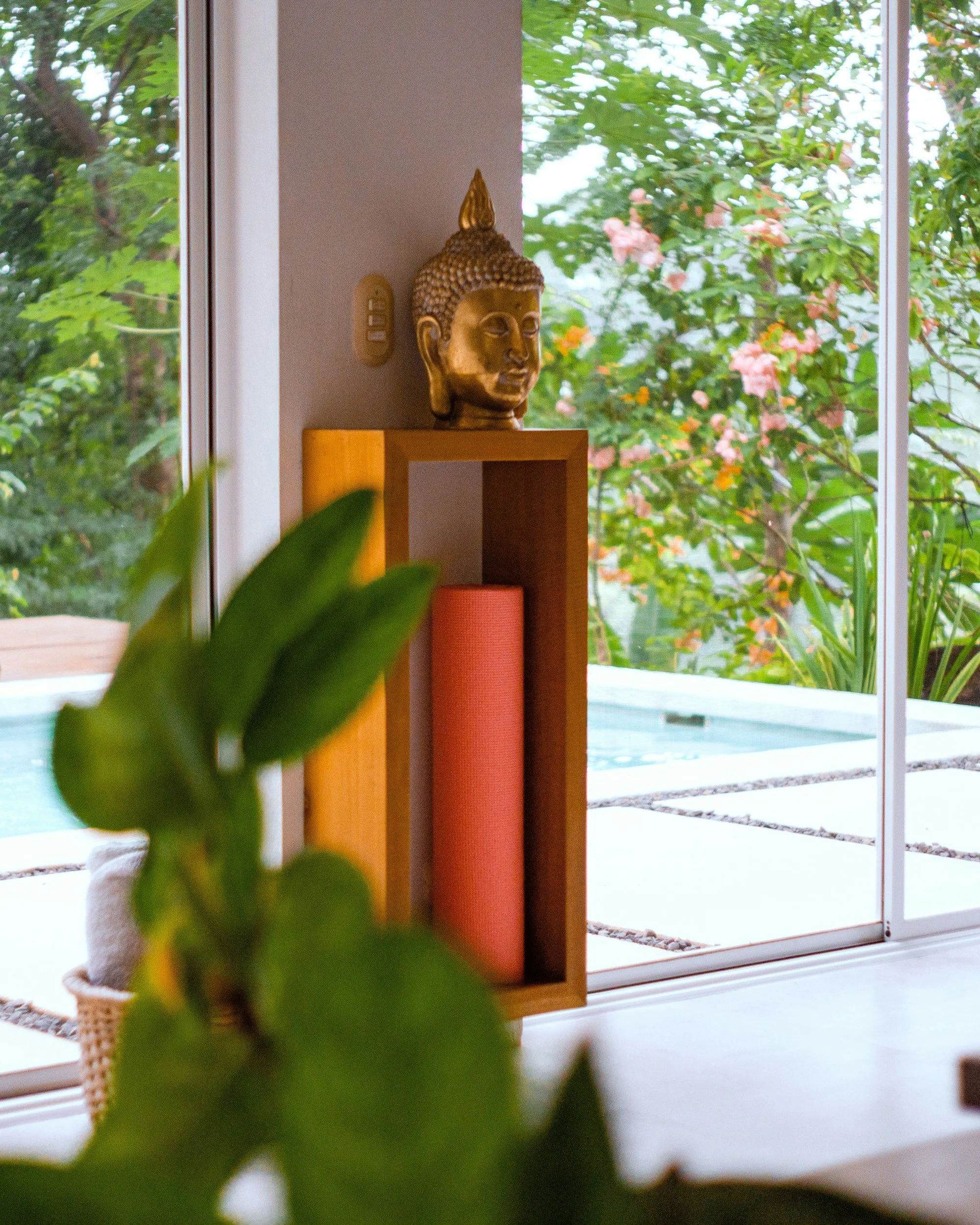 Indoor scene with a Buddha statue on a shelf near a window, with green foliage and pink flowers outside.