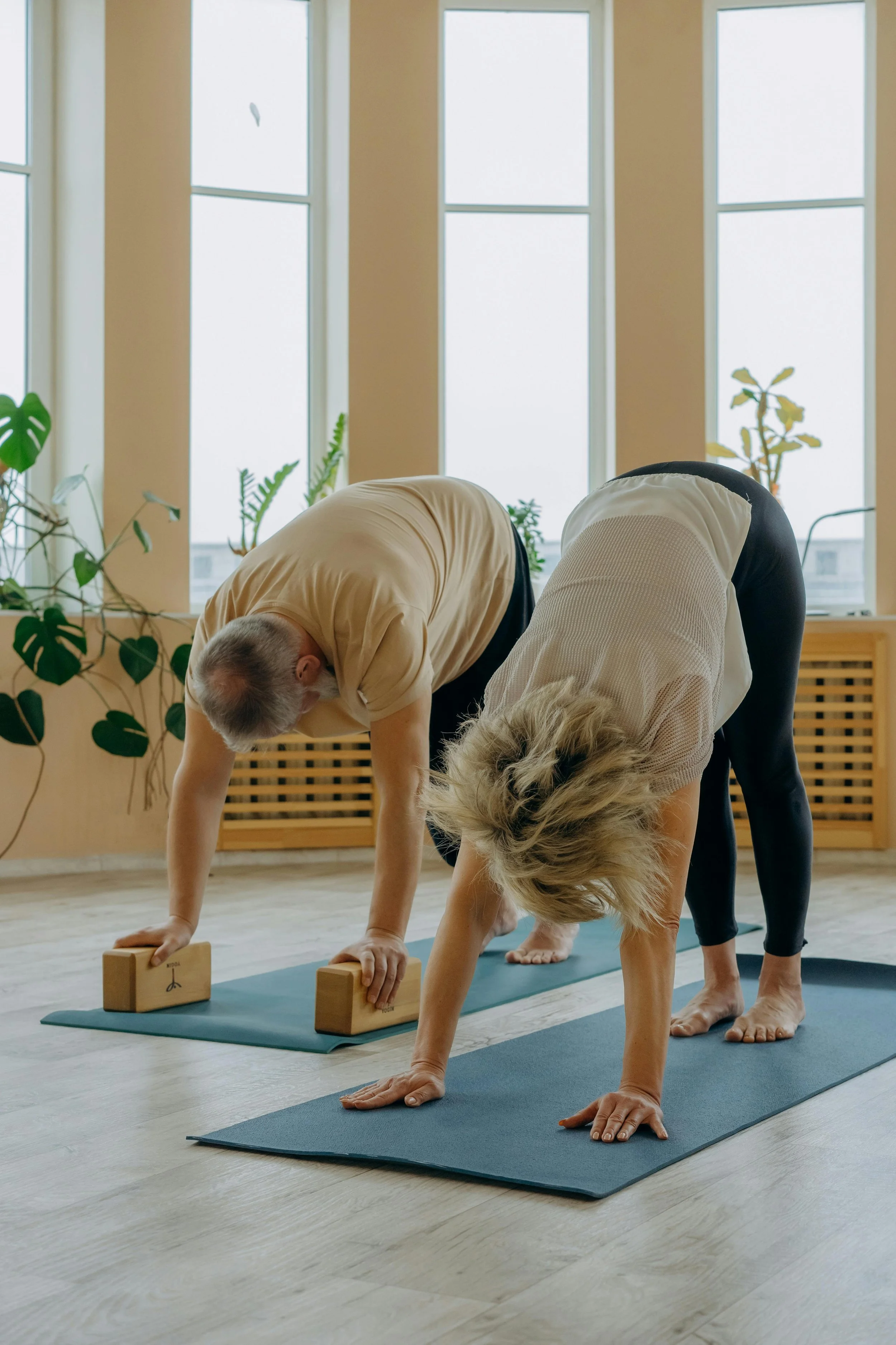 Two people practicing accessible yoga in a bright room with large windows, using yoga mats and yoga blocks.