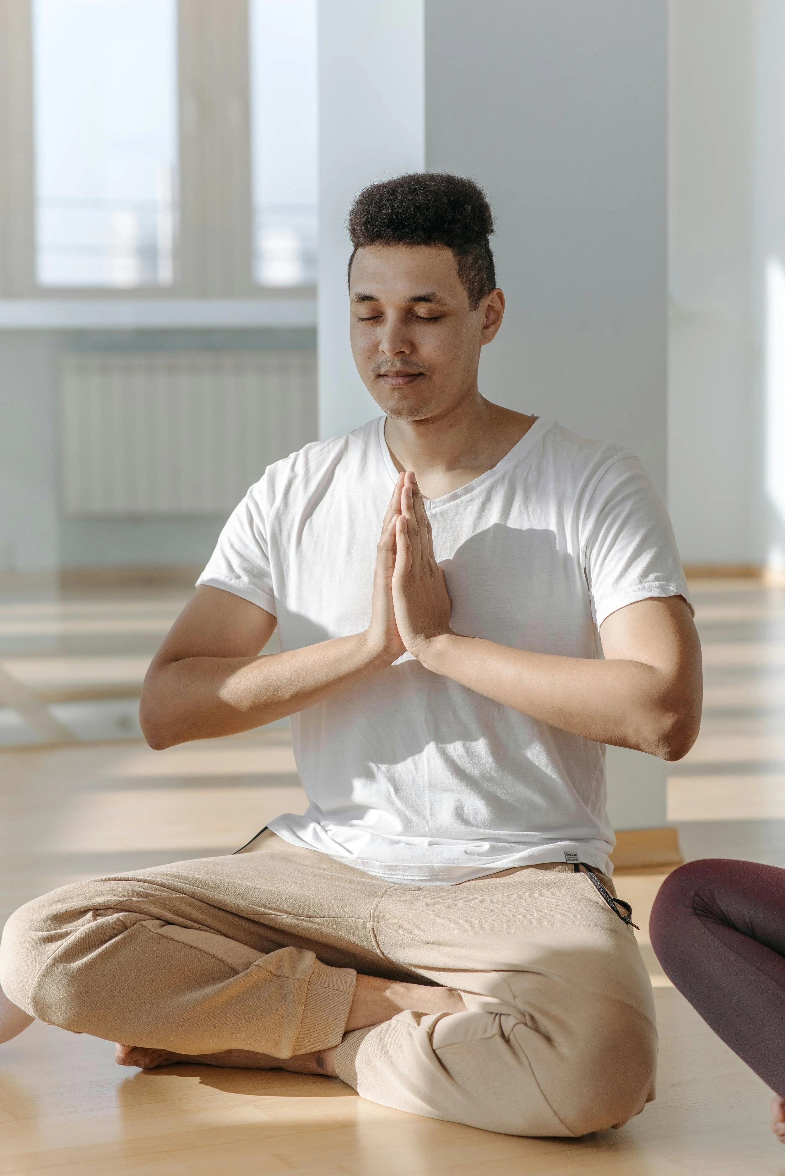 A man with short curly hair and wearing a white t-shirt and beige pants, sitting cross-legged on the floor with eyes closed and hands pressed together in a prayer or meditation pose in a bright, airy room.