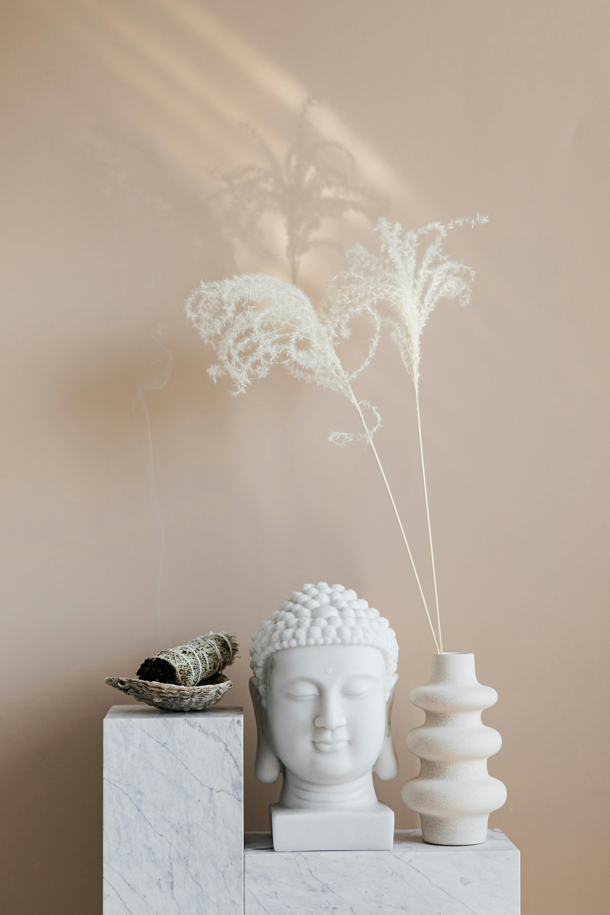 Decorative display featuring a white Buddha head sculpture, a tall white vase with dried branches, a bundle of sage on a marble pedestal, and a white textured sculpture in the background, arranged on a marble surface against a plain beige wall.