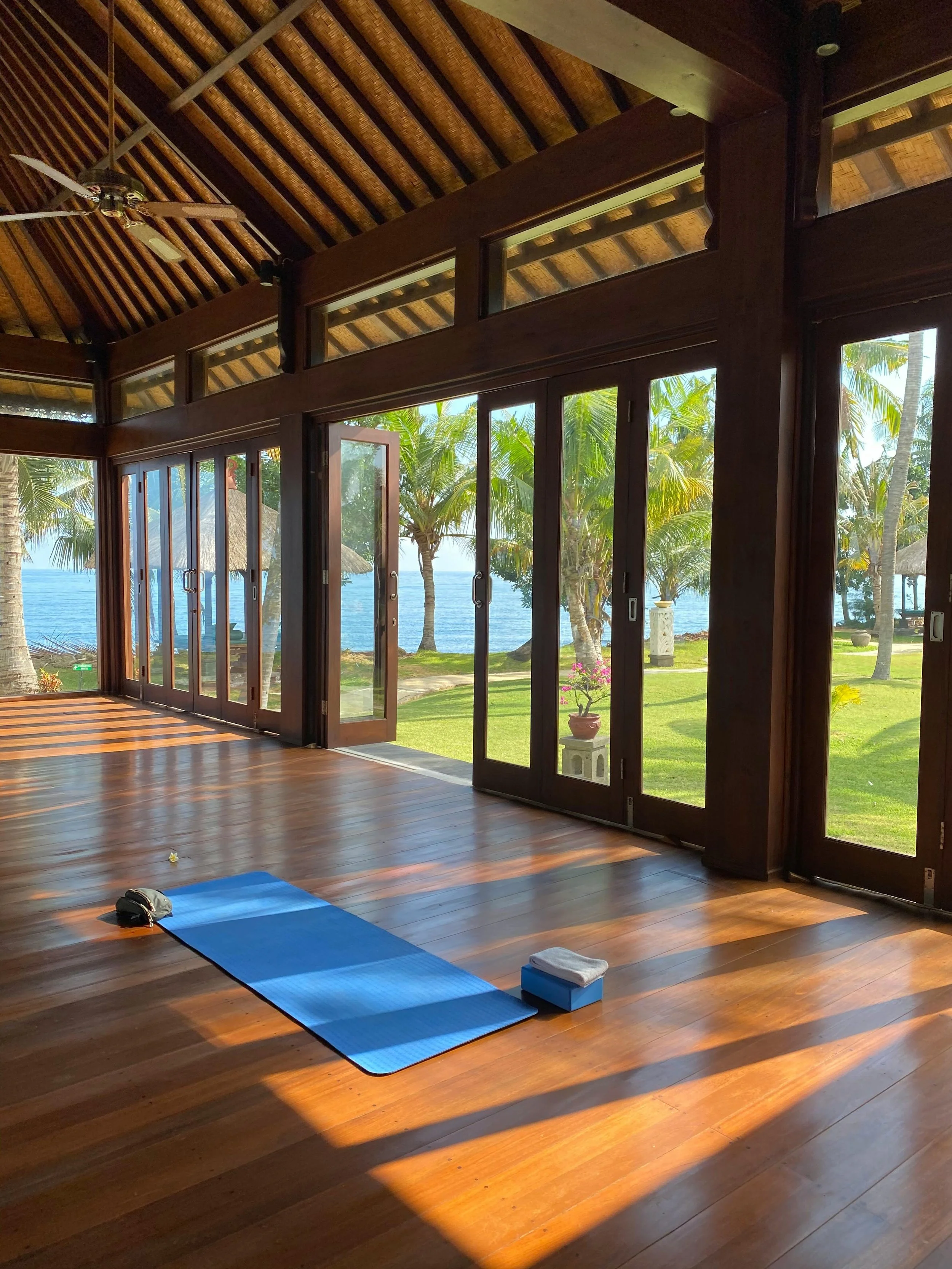 A yoga or meditation room with a blue mat on a wooden floor, a water bottle, and a white pillow. Large glass doors and windows open to a tropical outdoor scene with palm trees, grass, and water in the distance. The ceiling is made of wooden beams and thatch, with a ceiling fan.