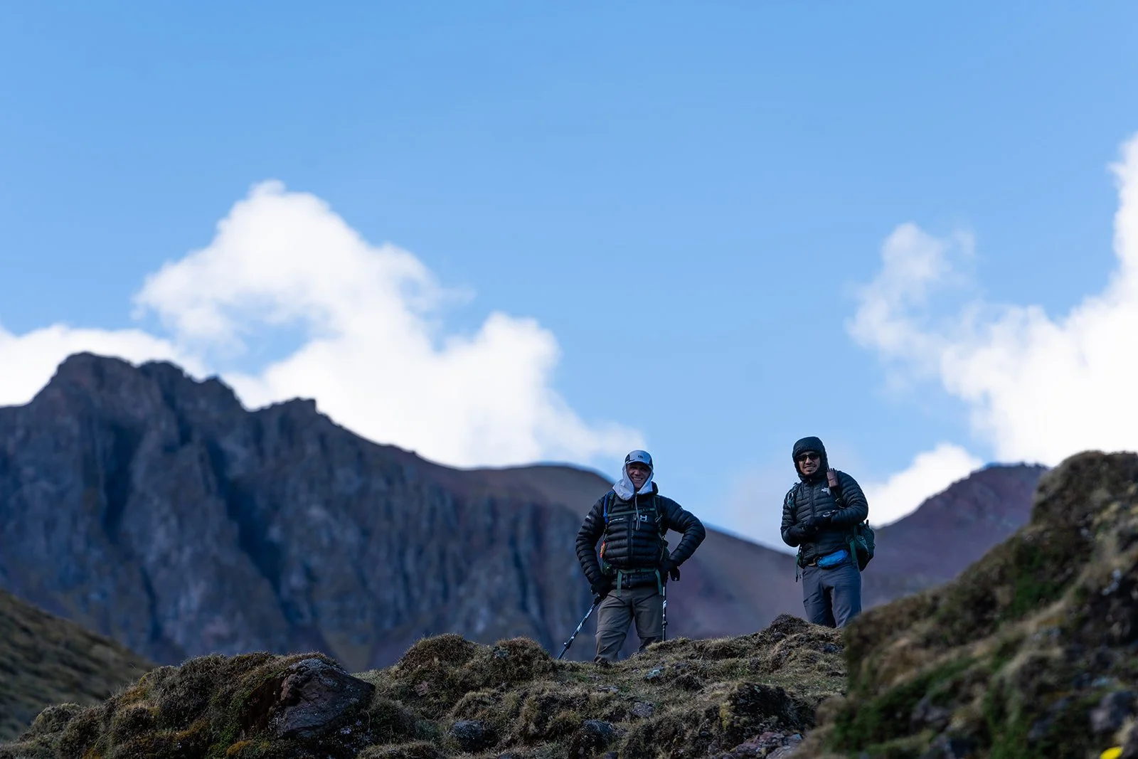 Two hikers standing on rocky terrain in a mountainous landscape, wearing outdoor gear and backpacks, with a mountain range and a partly cloudy sky in the background.