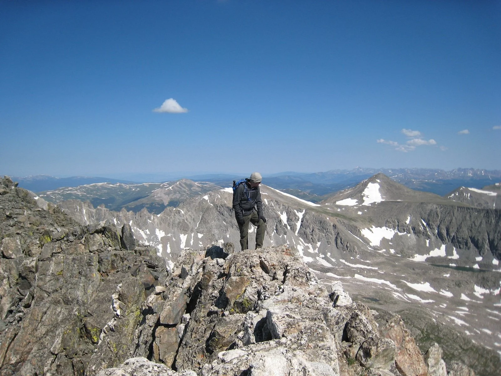 A person hiking on a rocky mountain trail with snow patches, surrounded by mountain ranges under a clear blue sky with a few clouds.