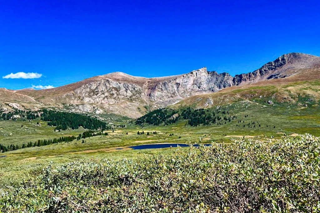 Scenic mountain landscape with green hills, a lake, and rocky peaks under a clear blue sky.