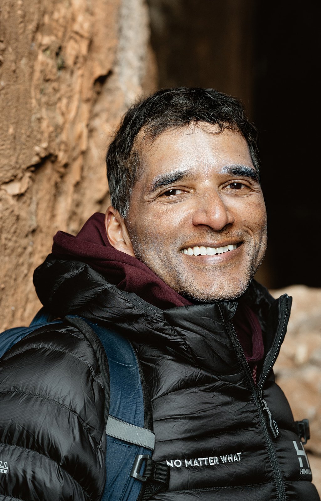 Smiling man in outdoor hiking gear, including a black puffy jacket, maroon hoodie, and a blue backpack, standing by a rocky wall.