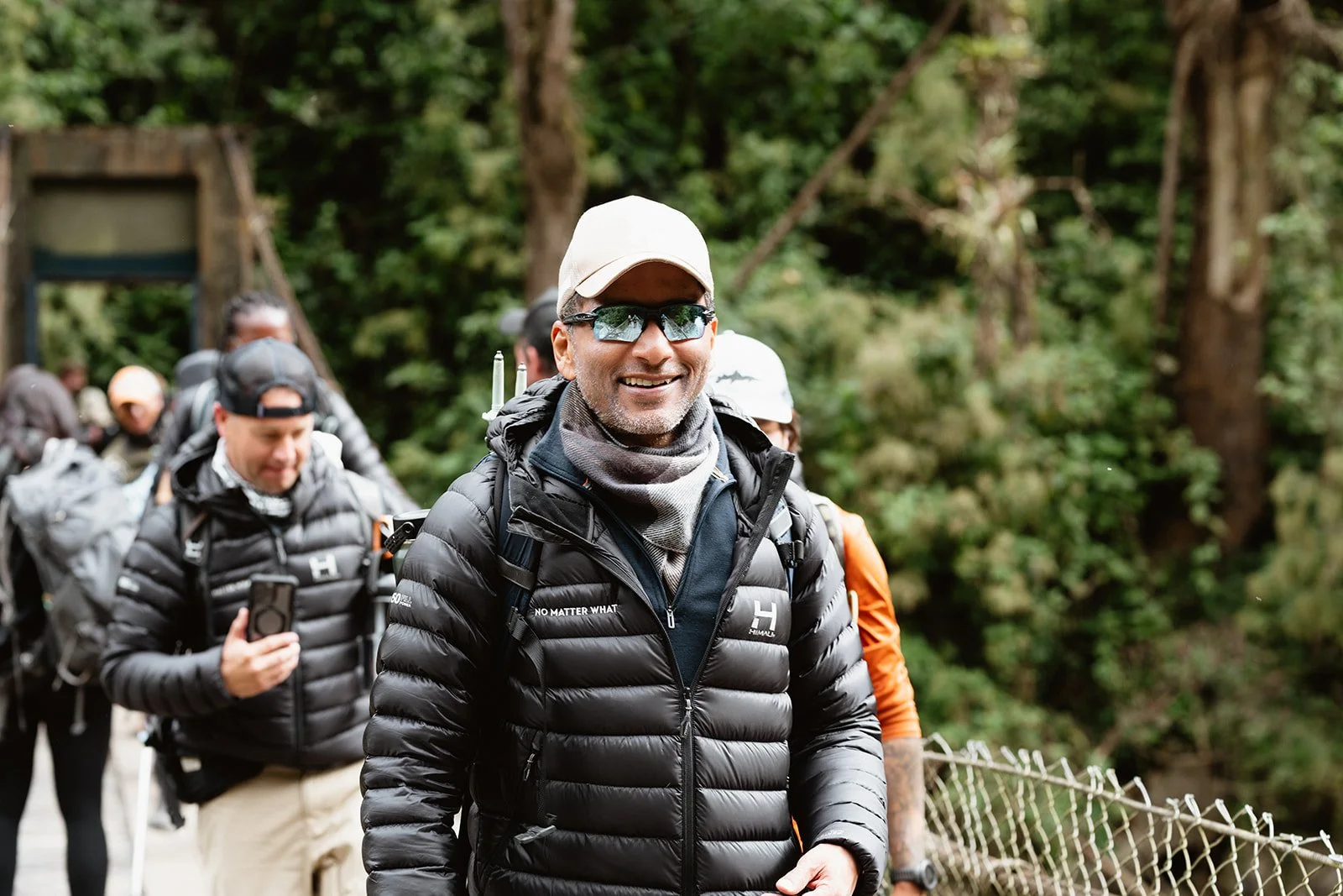 A group of hikers walking outdoors in a forest. The man in the front is smiling, wearing sunglasses, a beige cap, black jacket, and has a backpack. Other hikers are visible behind him, some taking photos, with trees and greenery around.