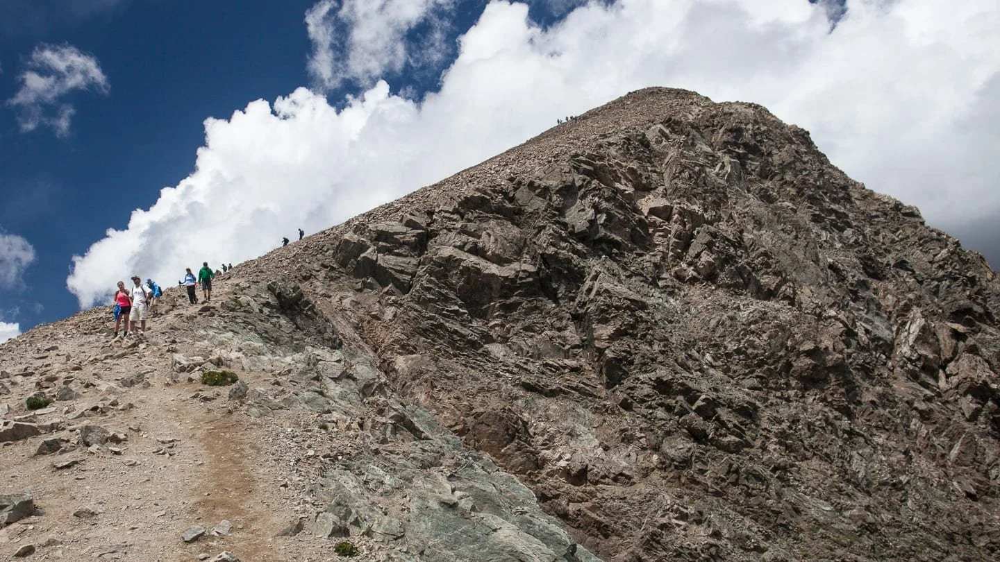 Group of hikers climbing a rocky mountain trail under a cloudy sky.