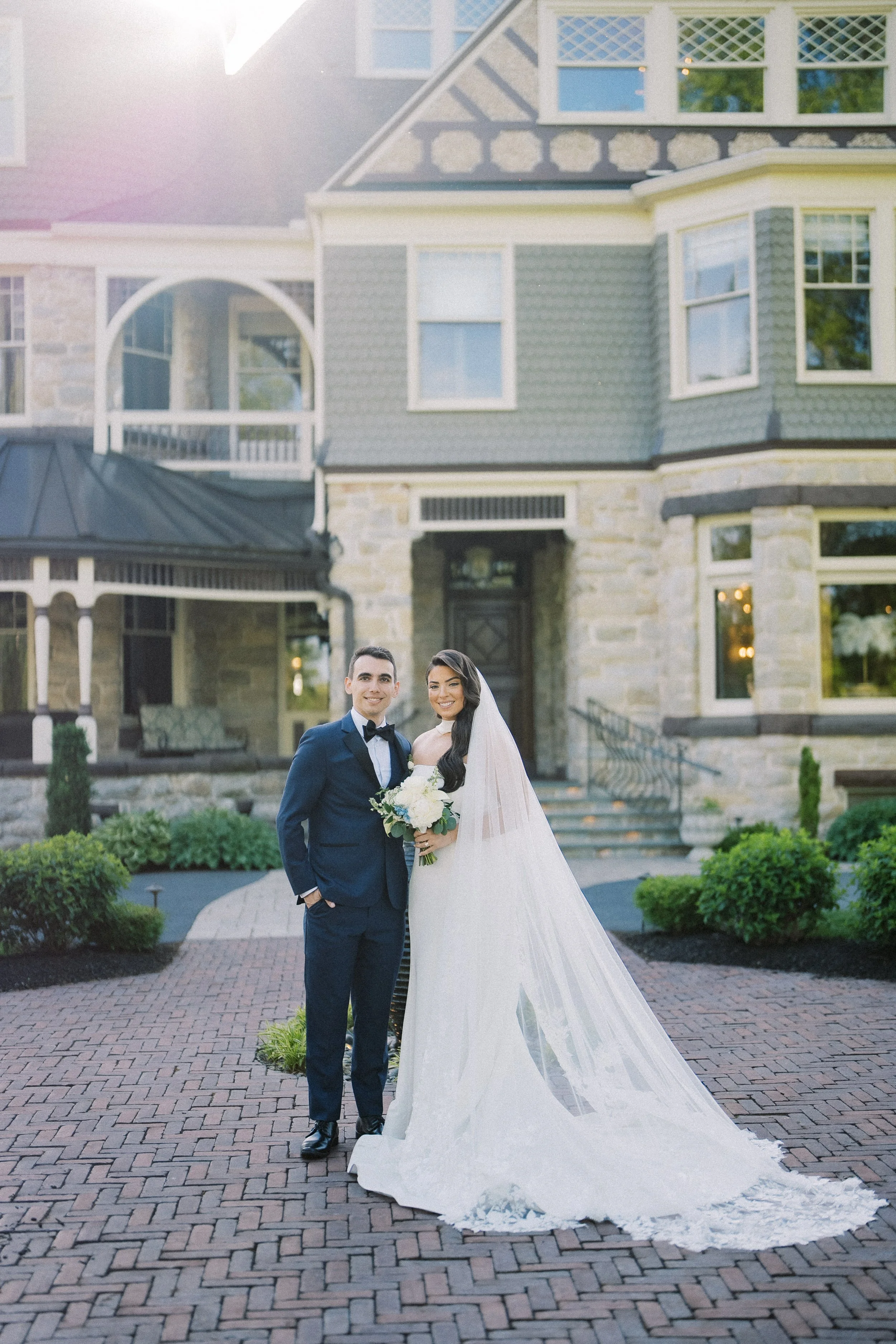 A newlywed couple stands outside a large stone and siding house. The groom wears a navy suit with a bow tie, and the bride wears a white wedding gown with a long veil, holding a bouquet of white flowers.