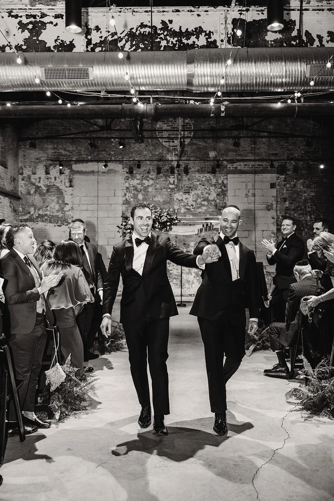 Two men in tuxedos holding hands and smiling, walking down an aisle at a wedding reception with guests clapping and celebrating.