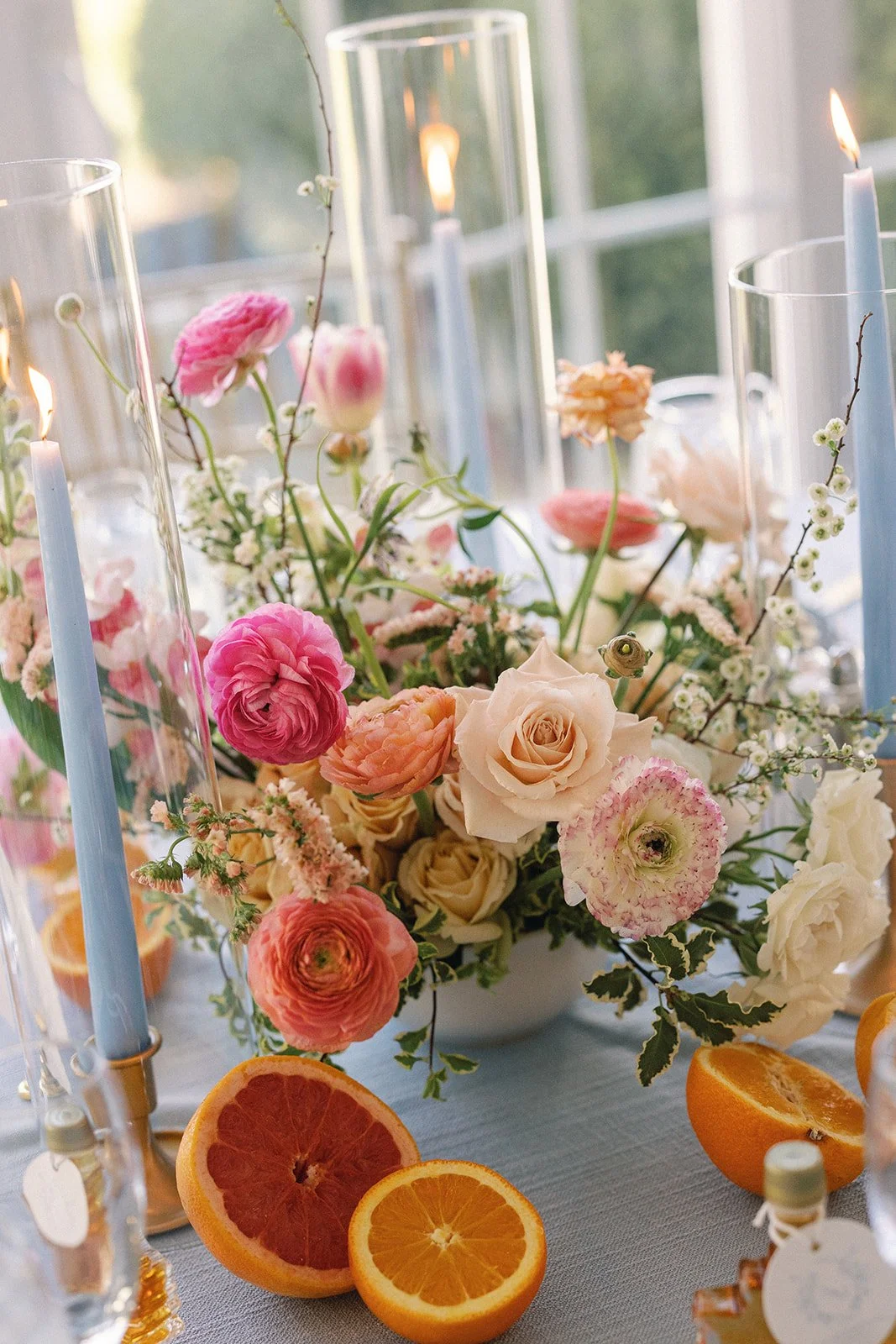 A floral centerpiece with pink, cream, and peach flowers on a table, surrounded by halved oranges and candles in tall glass holders.