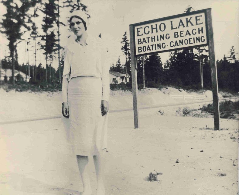1930. Inez Carson by the new Echo Lake sign
1930. A new sign replaced the old one. Houses in background: Burke, Monks, Henry Plate. Shows new road cut for Highway 99 and and the old brick road behind Firlands Way located at 195th and Aurora. Inez Car