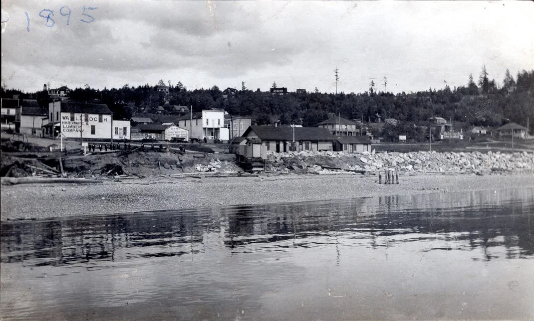 1890s. Richmond Beach business district seen from Puget Sound. 1907. The town fathers are standing at railroad station, possibly the opening day of the train depot. Sign of Walloch Hardware and Lumber. Cars parked on street. Photographer is standing 