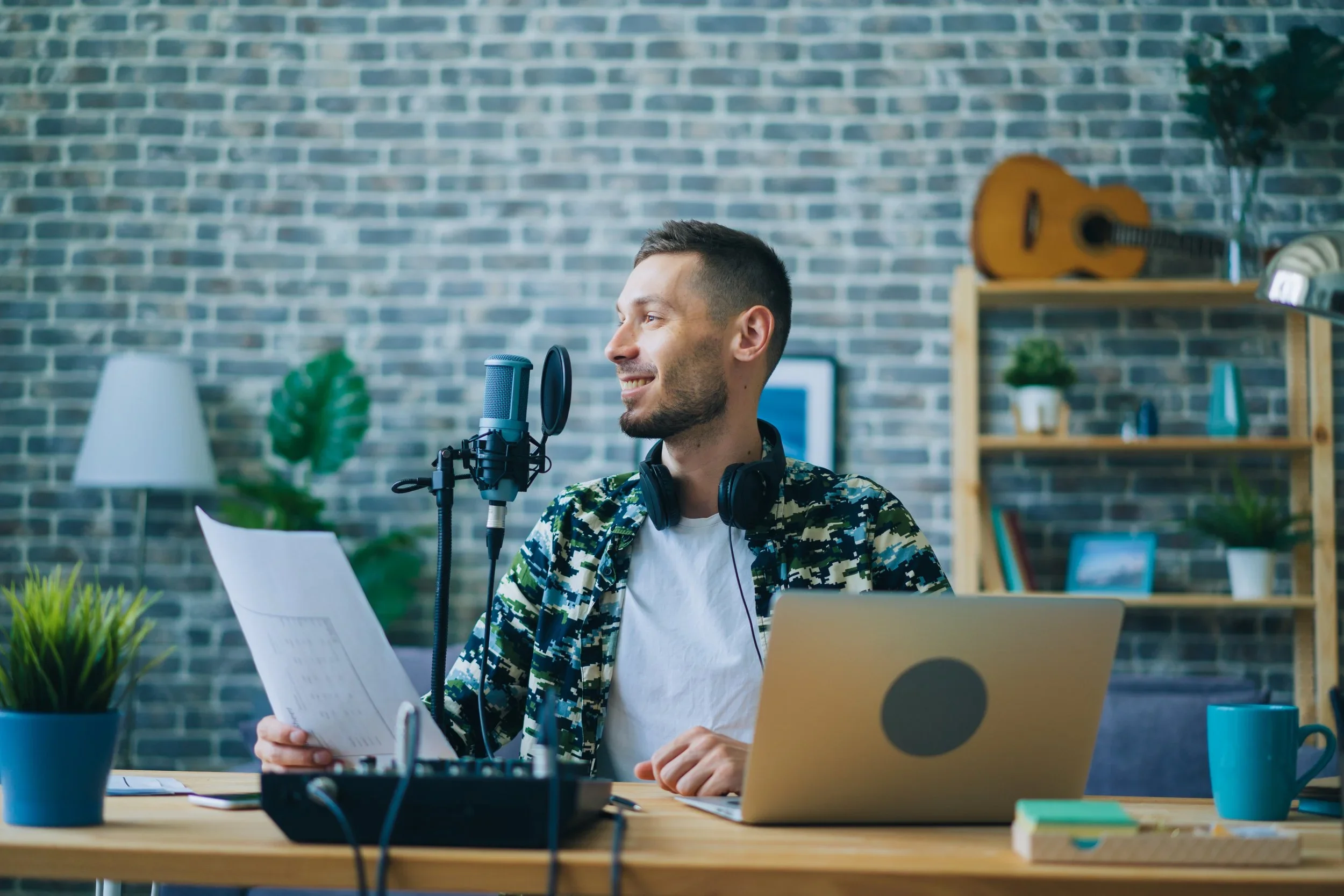 A man speaking into a microphone with headphones around his neck, sitting at a desk with a laptop, paper, and a coffee mug, in a room with a brick wall and shelves with books and decorative items. Podcasting in clematis street west palm beach