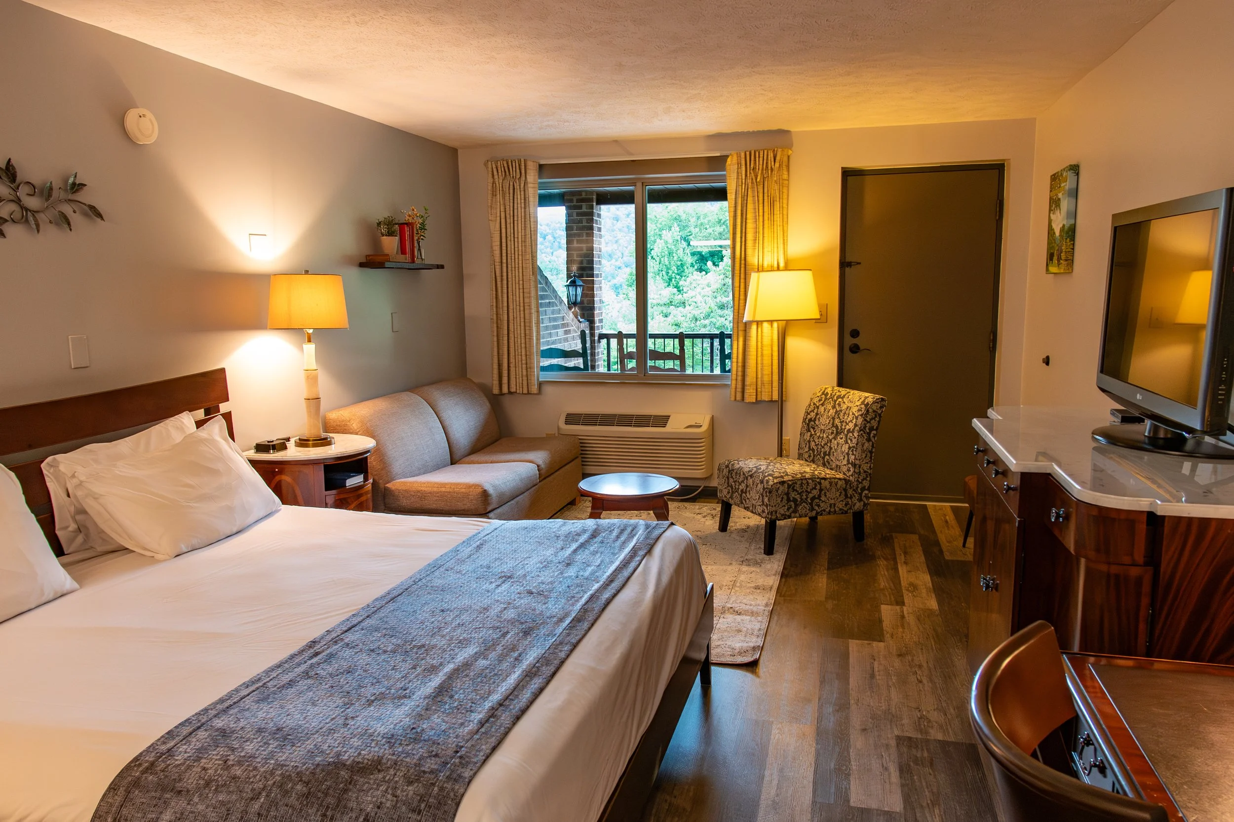 A hotel room with a bed, a beige sofa, a patterned chair, two lamps, a window with yellow curtains, a wall mirror, a TV on a wooden cabinet, and hardwood flooring.