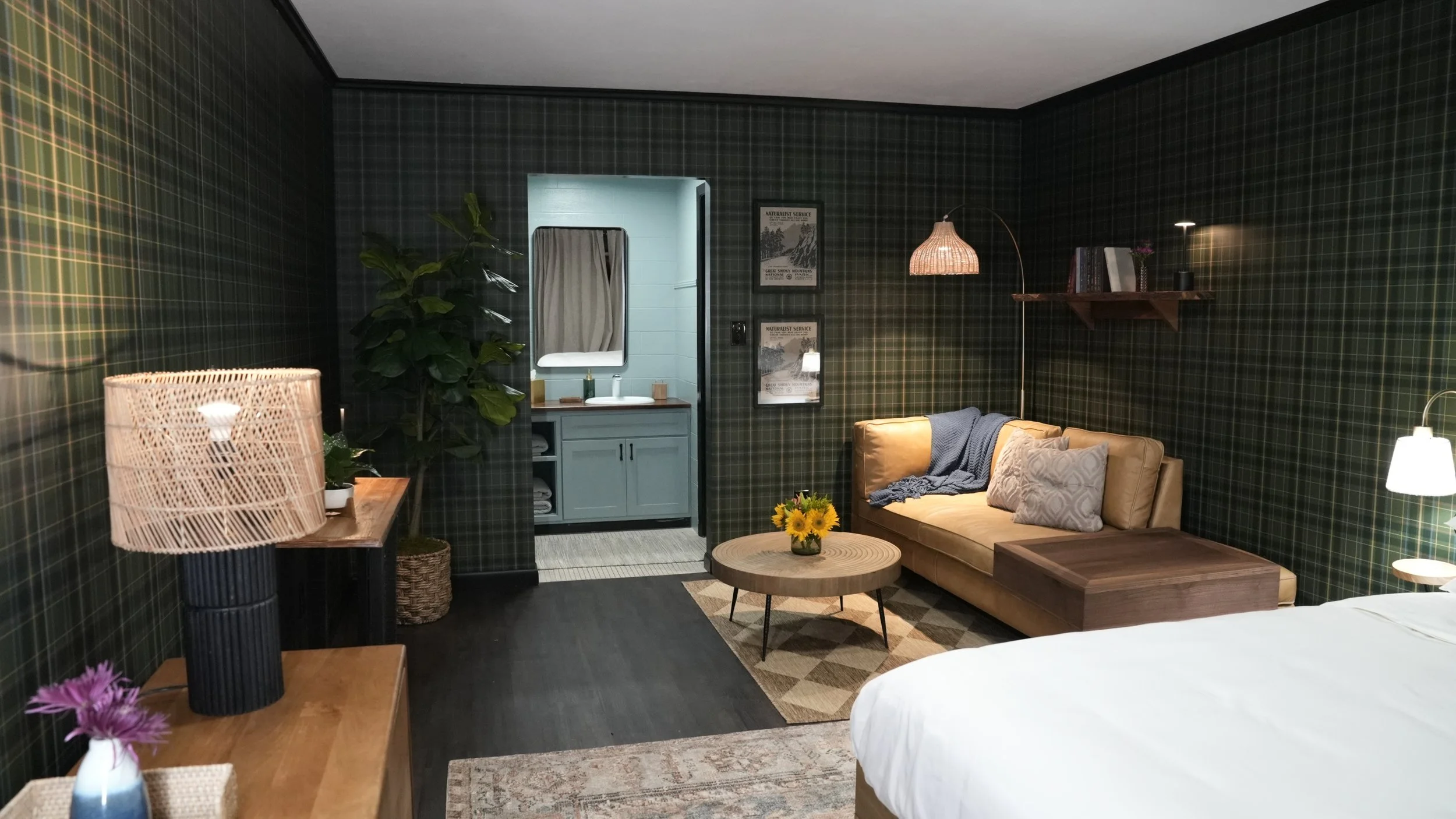 Cozy living area with dark plaid wallpaper, a beige sofa, a round wooden coffee table with a sunflower bouquet, a wall shelf, and a small bathroom visible through an open doorway.