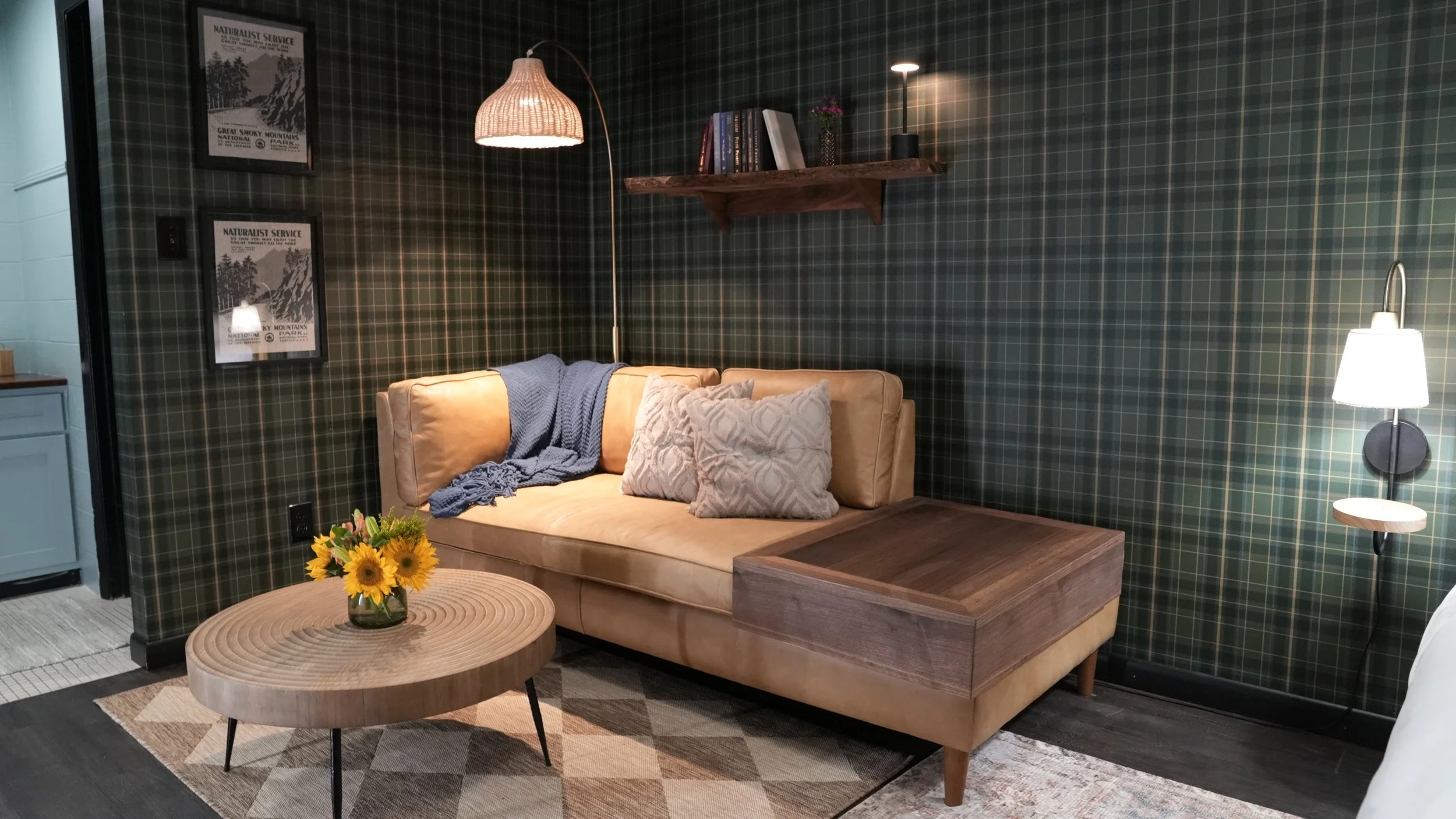 A cozy living room corner with a beige sofa holding a blue blanket and gray pillows. There's a small wooden coffee table with a sunflower arrangement, checkered wallpaper, a wall shelf with books and plants, a floor lamp, and framed posters on the wa