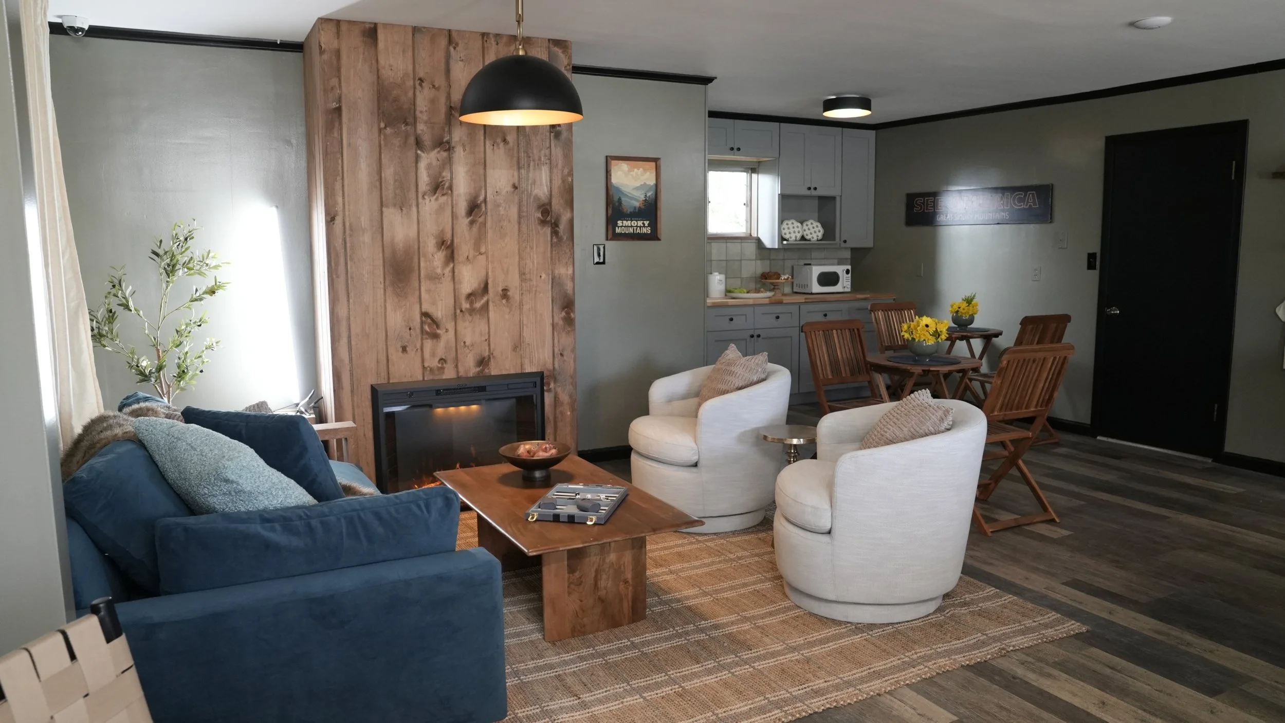 Living room with a blue sofa, two white armchairs, a wooden coffee table with a bowl and a game, a natural wood-paneled fireplace, a wooden dining table with four chairs, yellow flowers in two small vases, and a kitchen area in the background with gr