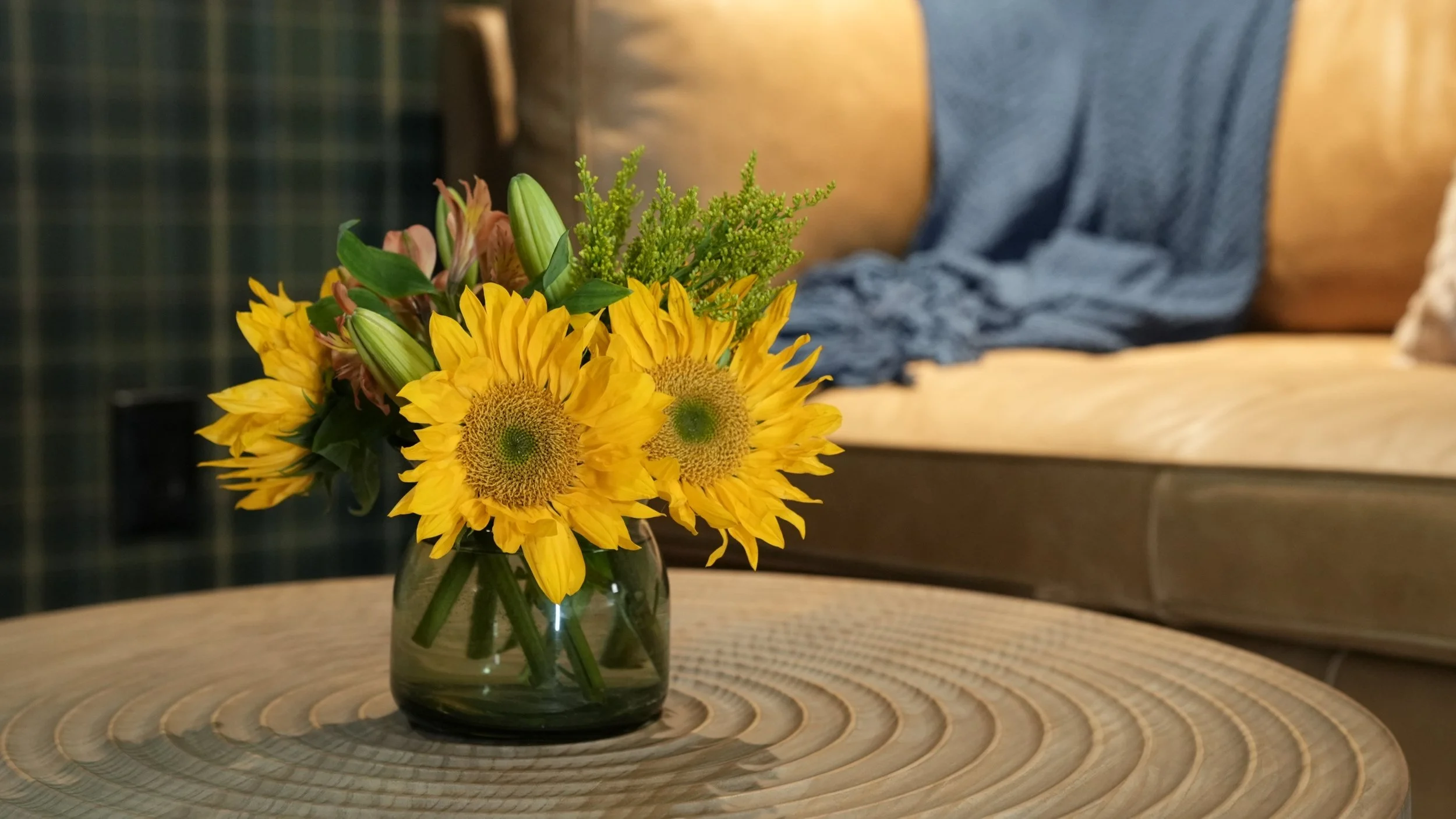A glass vase with a bouquet of yellow sunflowers, pink lilies, and green foliage on a round wooden table.