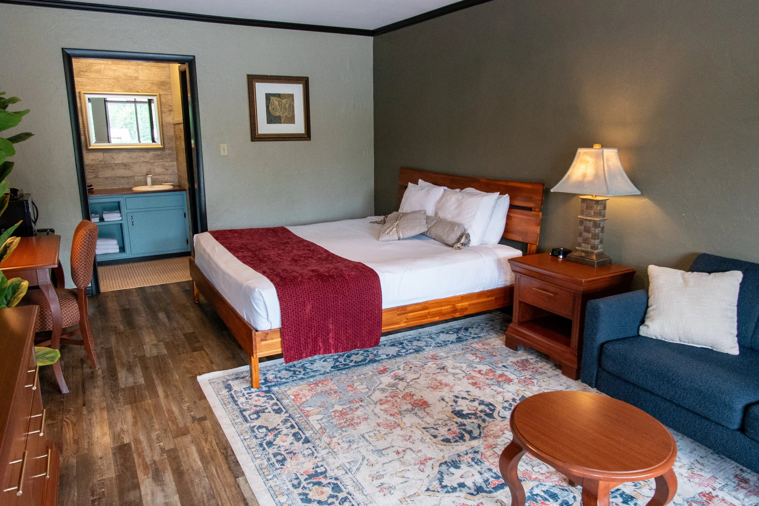 Hotel room with a bed, a blue sofa, a wooden nightstand with a lamp, a small wooden table, a colorful rug, and an adjoining bathroom with a sink, mirror, and window.