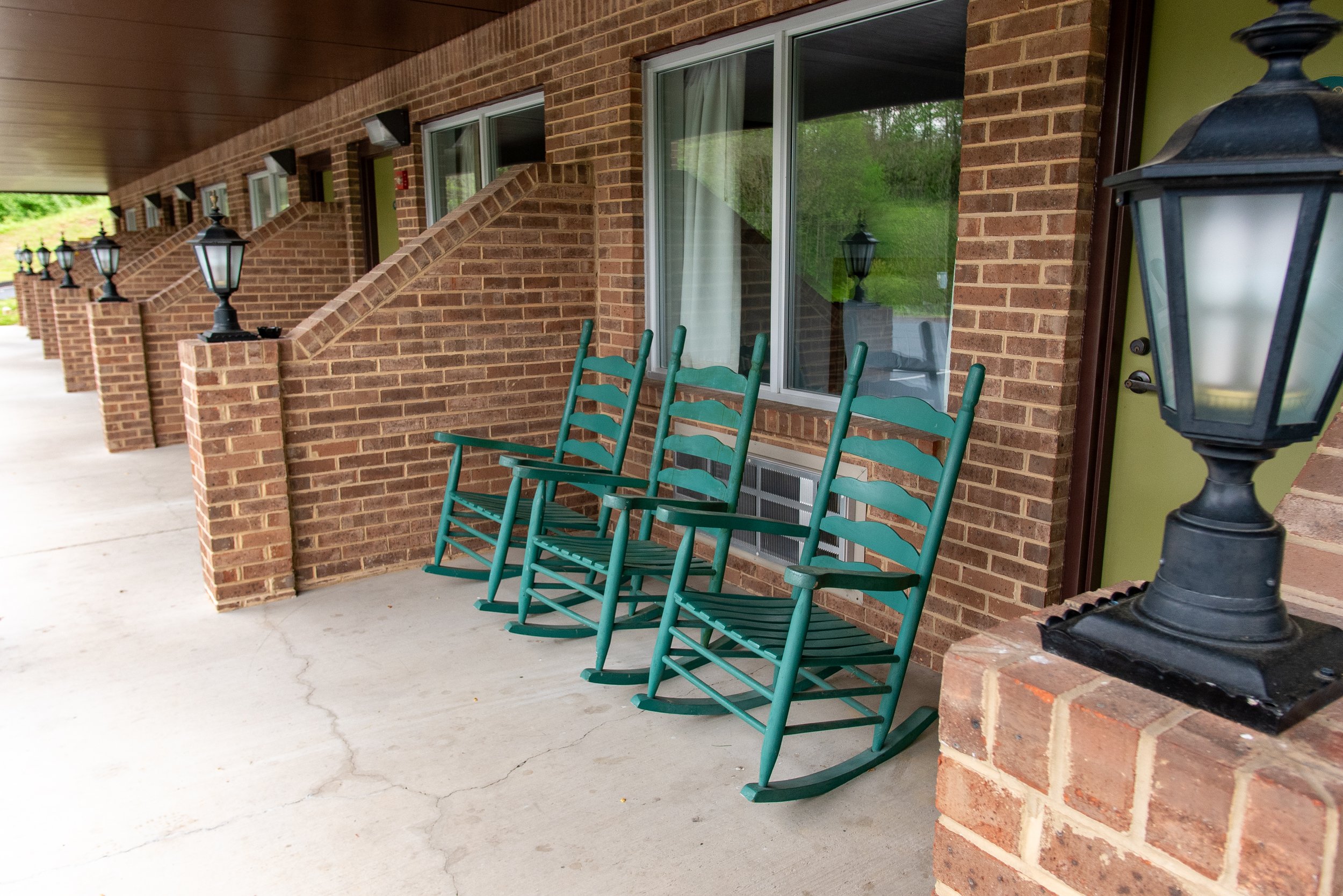 Four teal rocking chairs on a concrete porch next to a brick wall with large windows and black lantern-style lights.