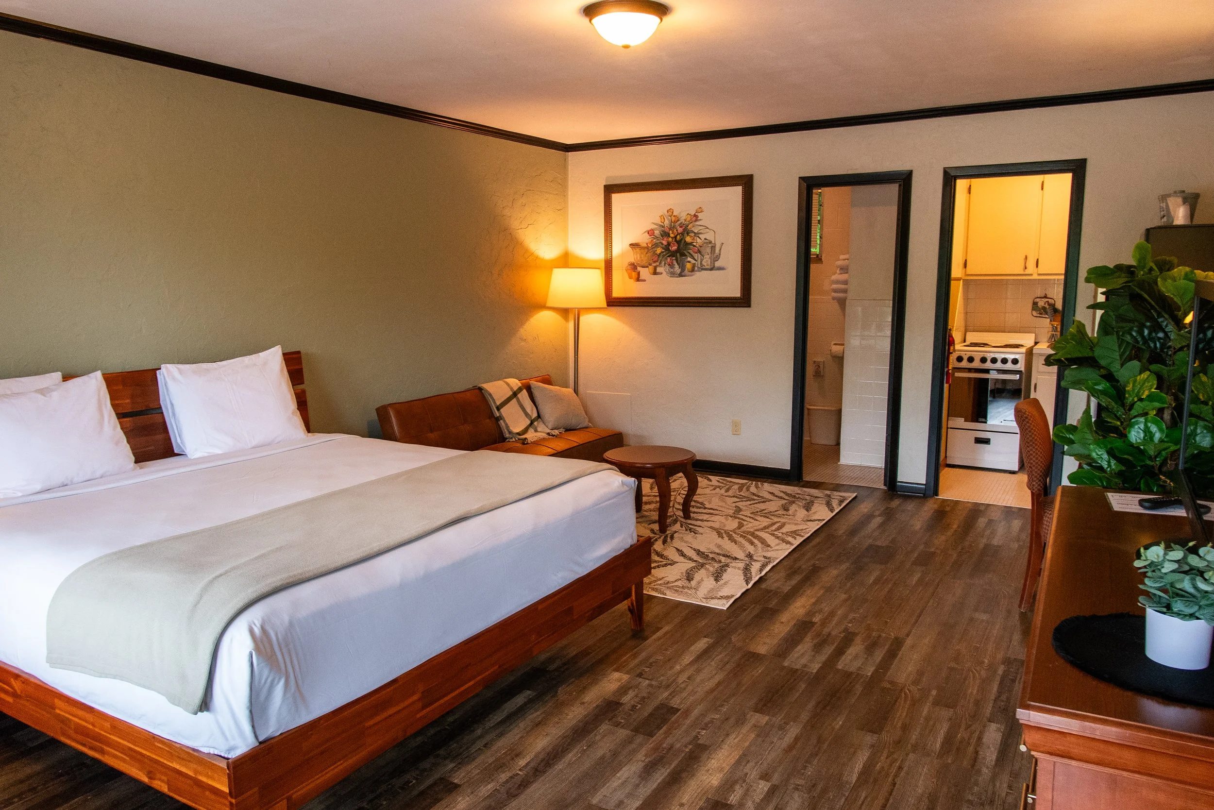 A hotel room with a large bed, a sitting area with a lamp, a painting on the wall, a door leading to a bathroom and a small kitchen, wooden flooring, and a dresser with a potted plant.