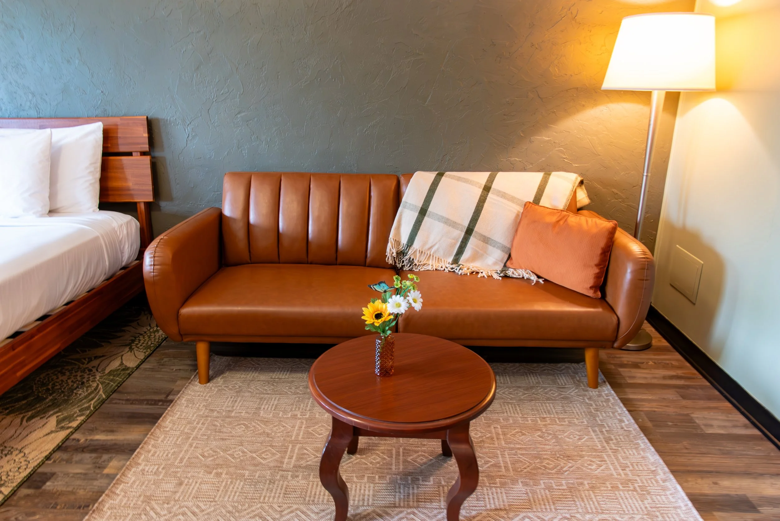 Living room with brown leather sofa, wooden coffee table with a vase of flowers, a bed with white linens, a floor lamp, a blanket, and an orange pillow, with hardwood and patterned rugs.