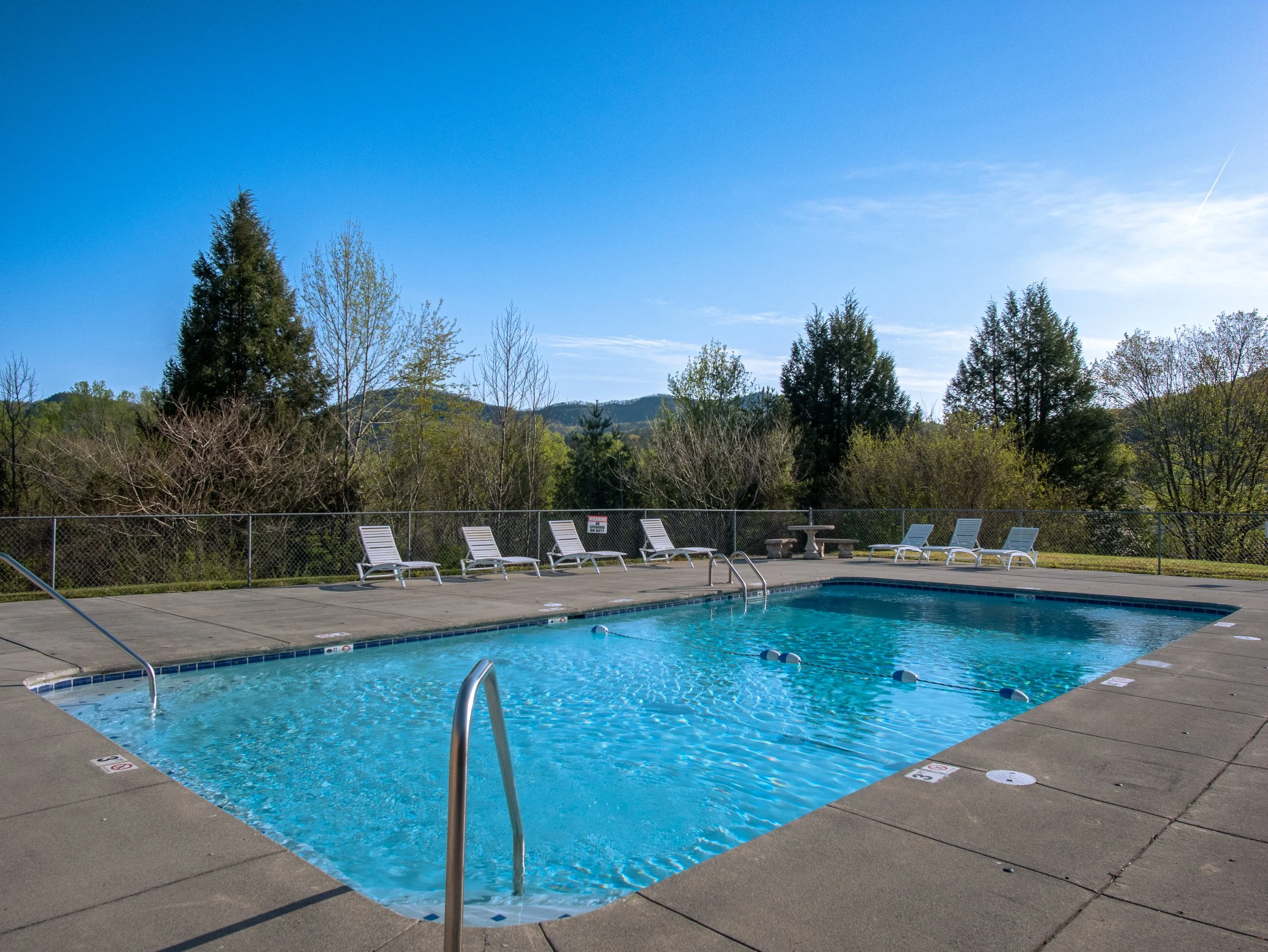 Empty outdoor swimming pool with metal handrails, surrounded by poolside chairs and scenic trees on a clear sunny day.