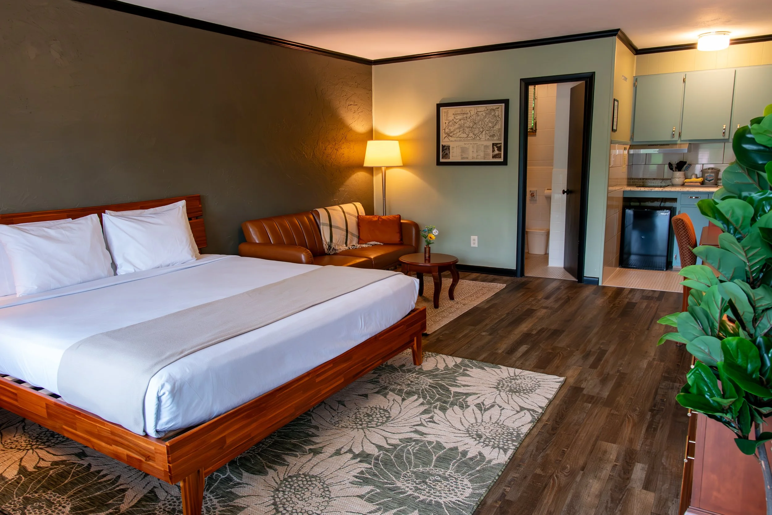 Hotel room with a bed, leather sofa, side table with flowers, lamp, framed map, small kitchenette, and door to bathroom, decorated in warm earth tones.