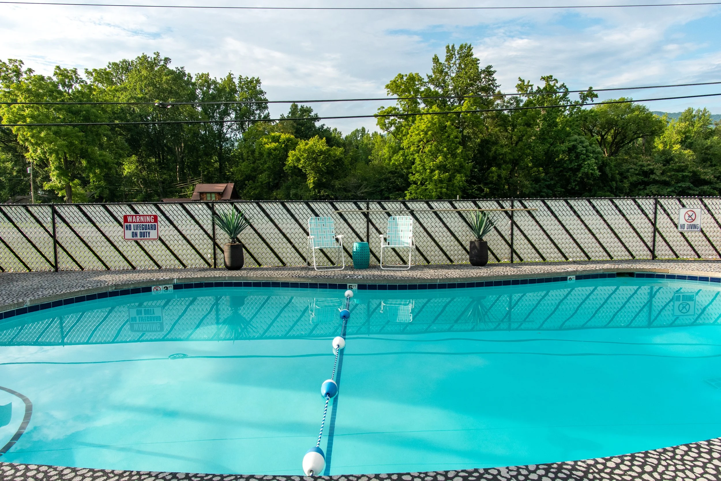 Swimming pool with two white lounge chairs, potted plants, and safety signs, surrounded by green trees and fencing.