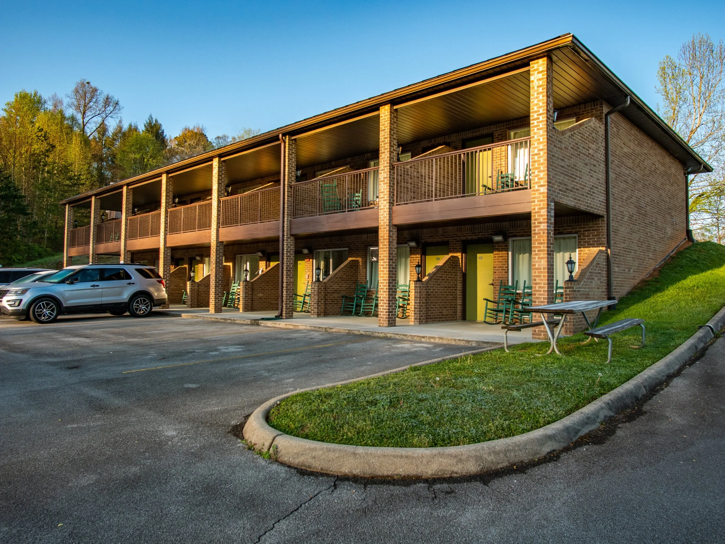 A two-story brick motel building with a parking lot in front. The building has a balcony on the second floor and yellow doors on the ground floor. Several cars are parked, and there is a bench and picnic table outside.