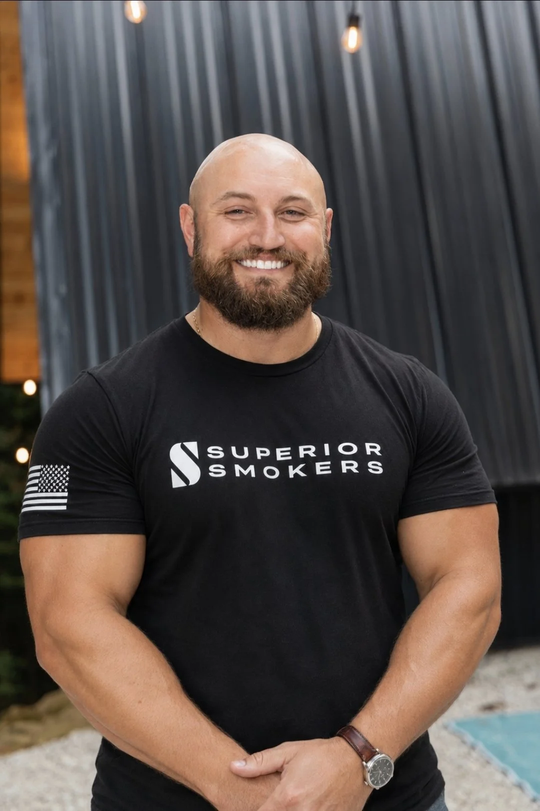 Jeremy Hengy, co-owner of Superior Smokers, smiling outdoors wearing a black Superior Smokers t-shirt. Modern building and outdoor string lights in the background