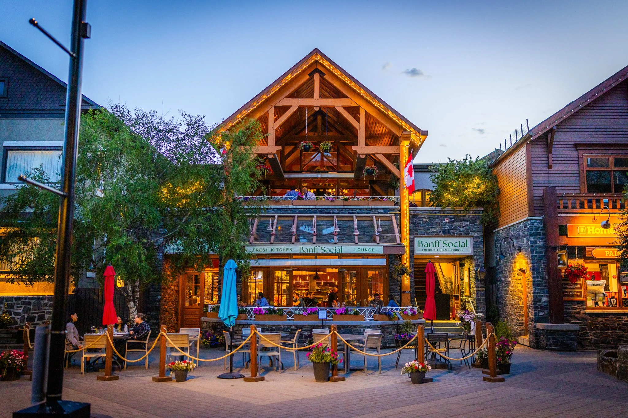 Outdoor view of a restaurant called Banff Social with a rustic wooden upper deck, string lights, and outdoor seating area, in a mountain town during evening.