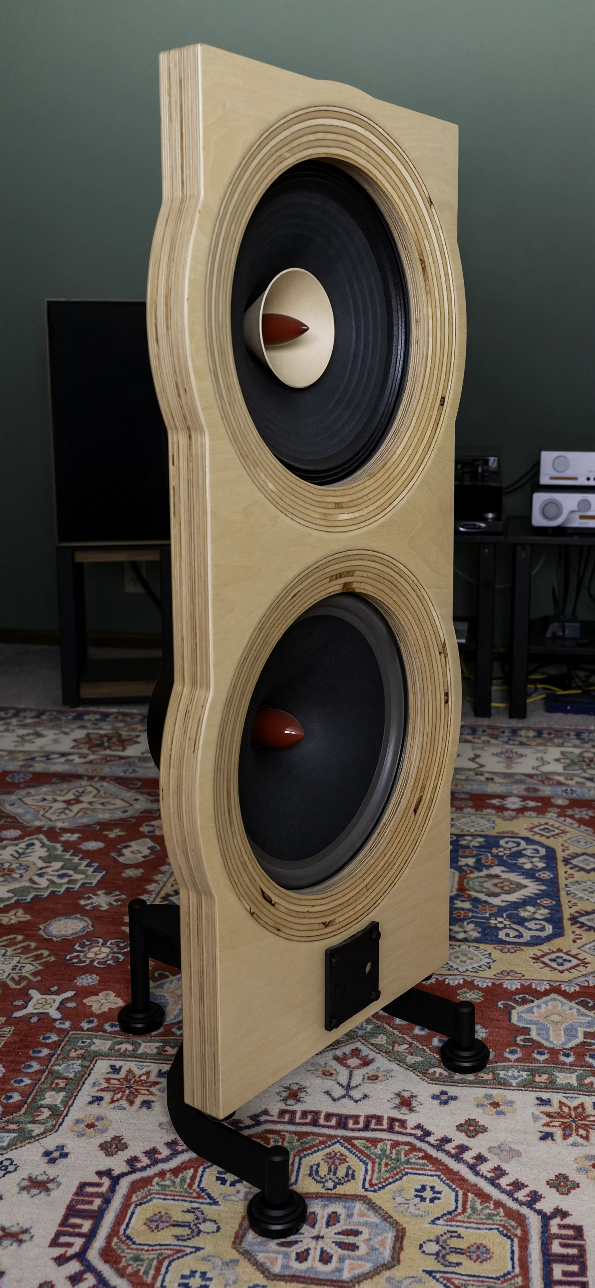 Three-quarter view of a Maco Macarias loudspeaker showing its sculpted Baltic birch baffle, exposed plywood layers, and open-baffle construction.