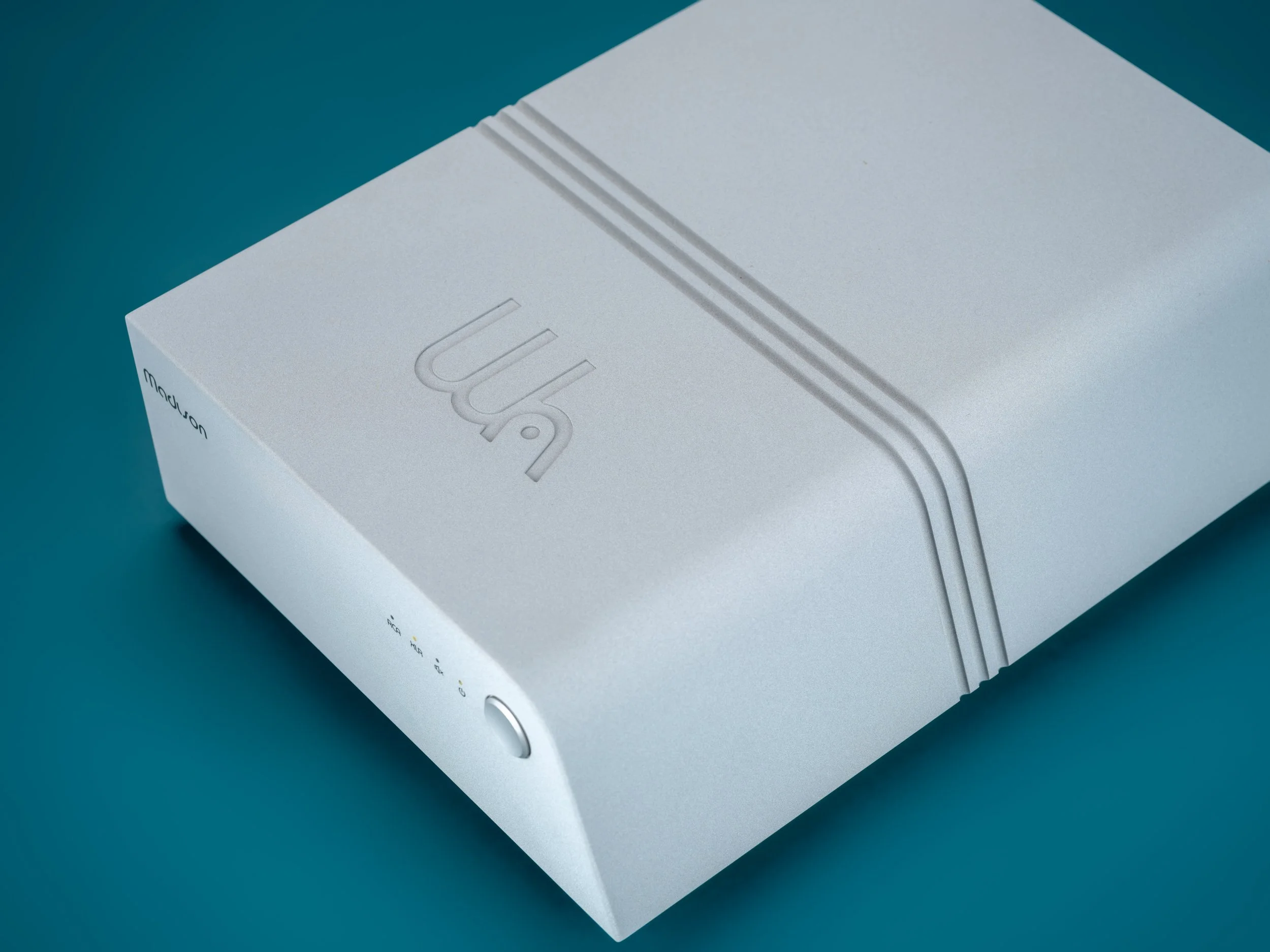 Wattson Madison Amplifier on a teal background, shown in an elevated three-quarter view highlighting its compact, machined aluminum chassis.