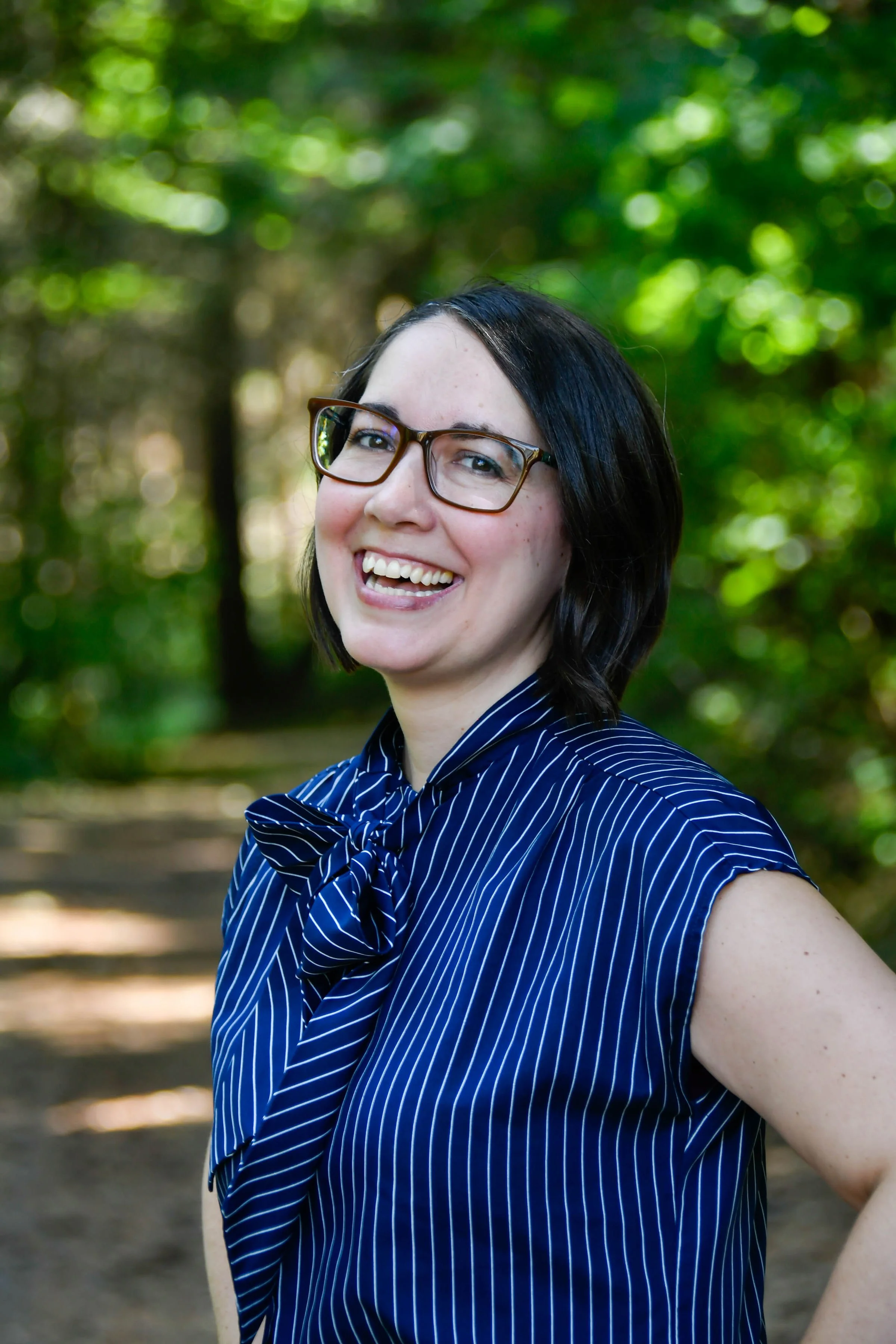 Photo of woman with light skin and dark hair with large smile in the foreground. She's wearing a vintage navy blue pin-striped shirt with large bow and dark rimmed glasses. There are green fir trees blurred out in the background.