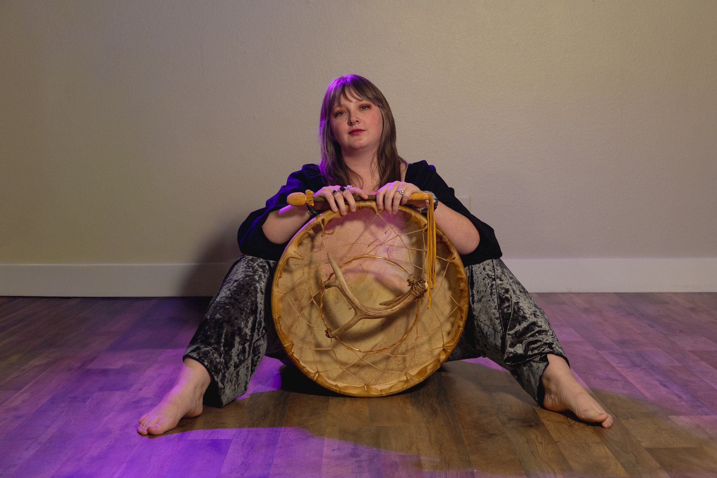 A woman with light brown hair sitting on a wooden floor against a plain beige wall, holding a drum with a wooden frame and skin surface, with a small antler attached to the drum. She is wearing patterned pants and a black top.
