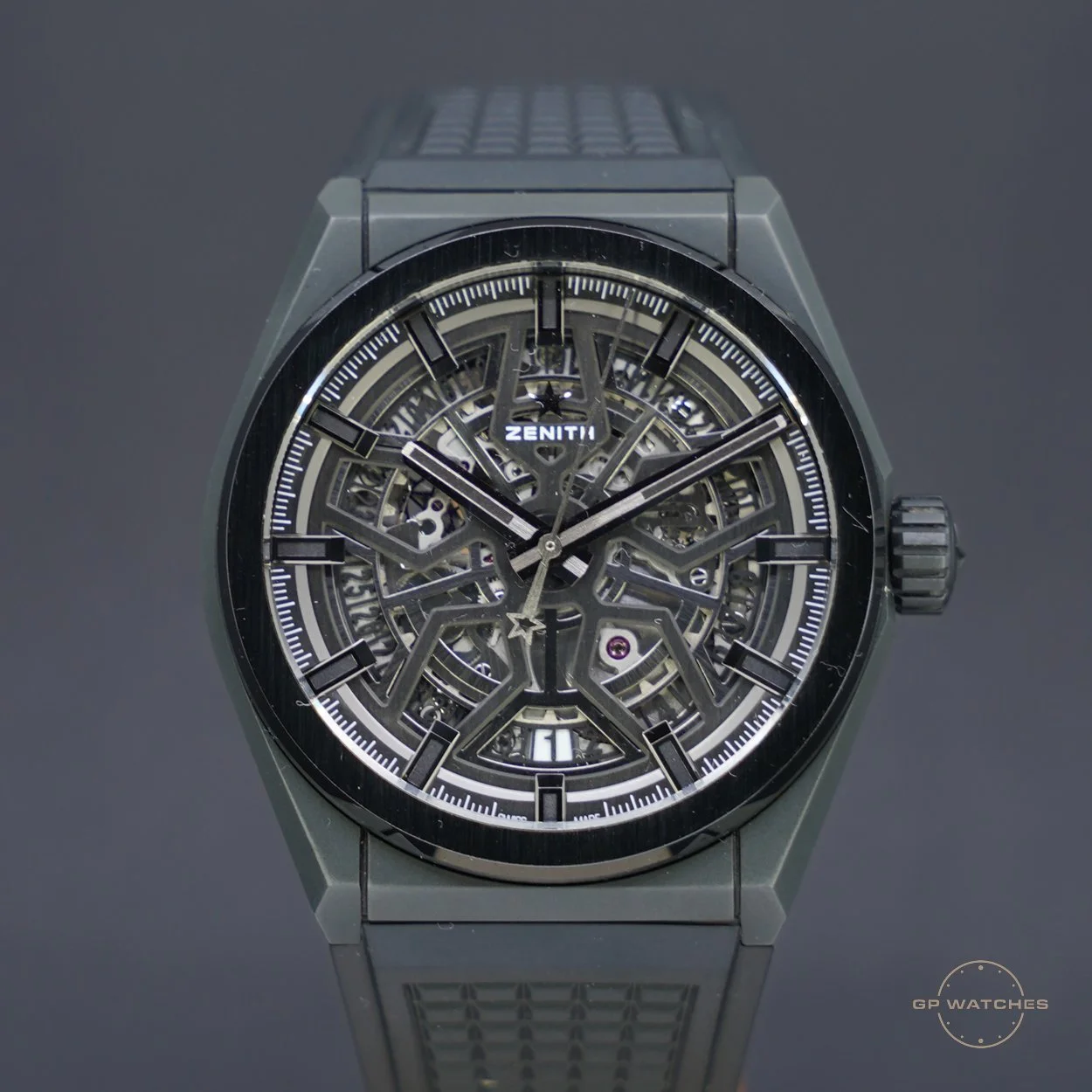 A luxury wristwatch with a skeleton dial revealing its mechanical movement, branded 'Zenith' with black hour markers, hands, and a date window at 6 o'clock, set against a dark background.