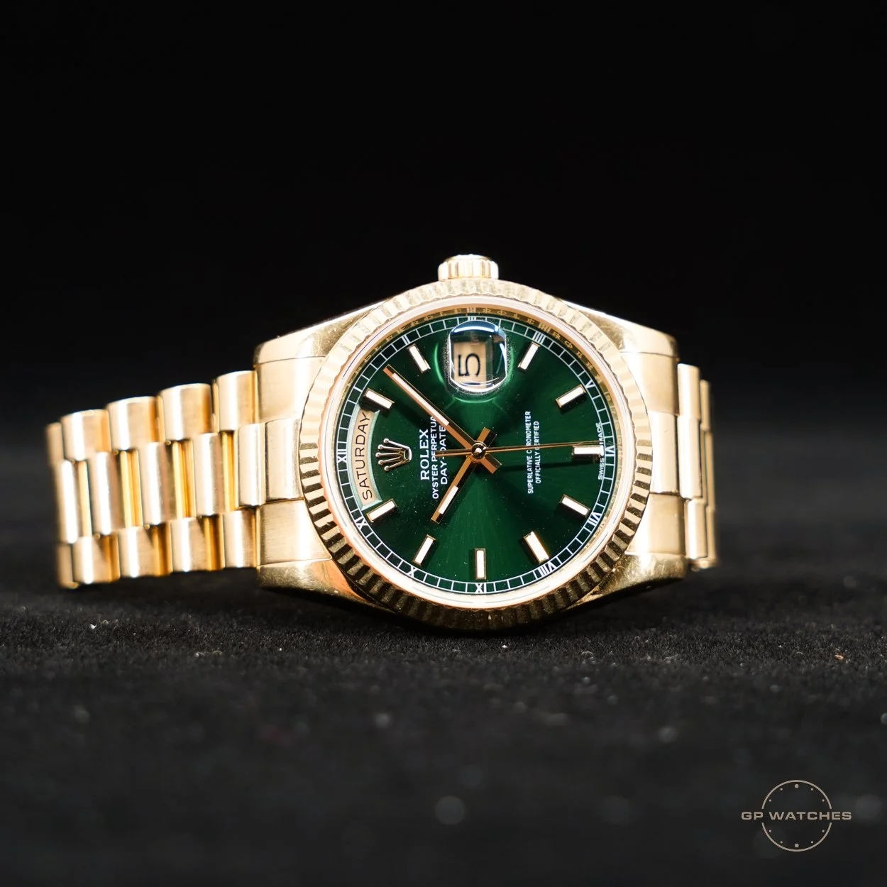 A gold Rolex watch with a green dial, date display, day of the week, and Roman numeral hour markers, set against a black background.