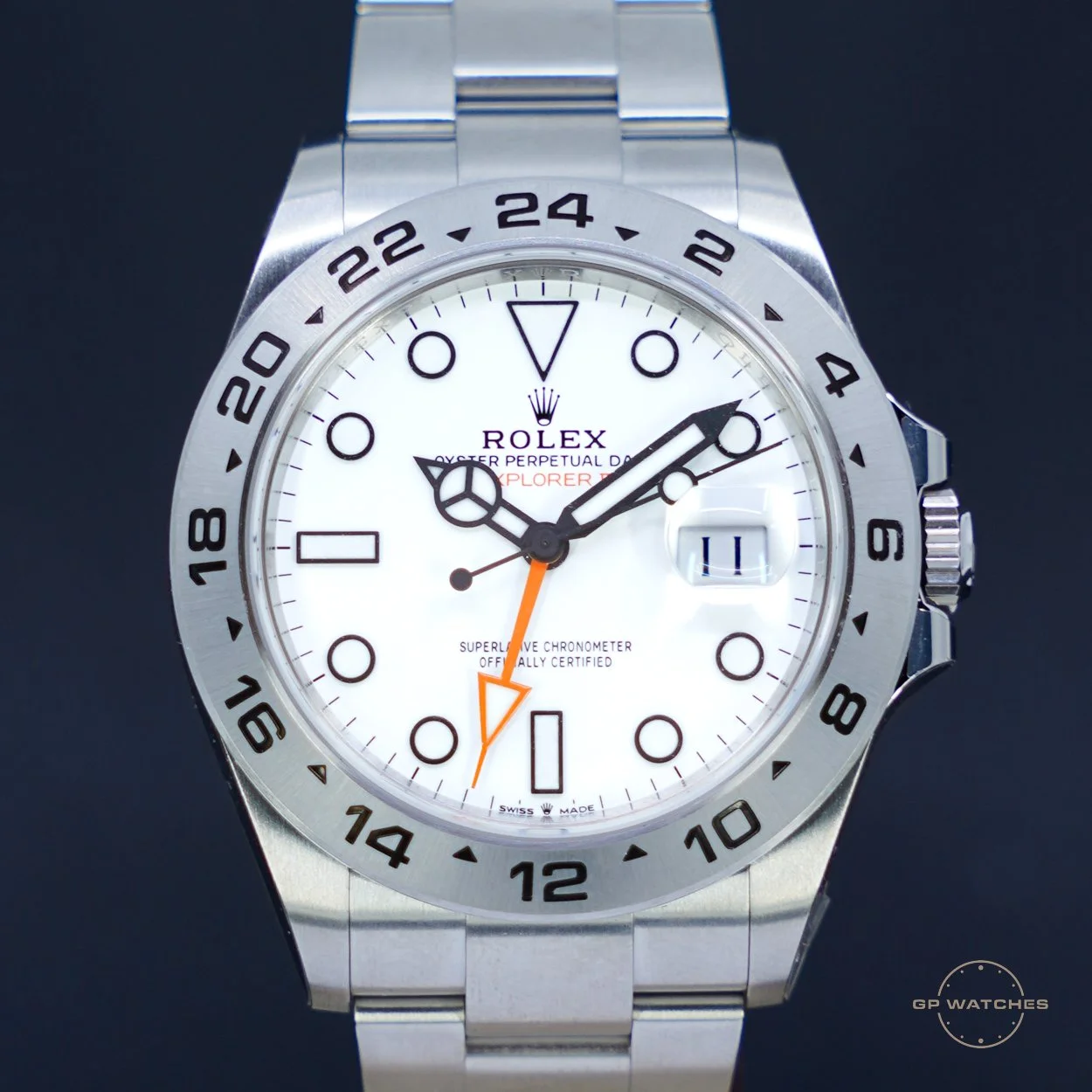 A stainless steel Rolex Explorer II watch with a white dial, black hour markers, black hands, a date window at 3 o'clock, and a black 24-hour bezel. The watch has black and orange accents on the second time zone hand.