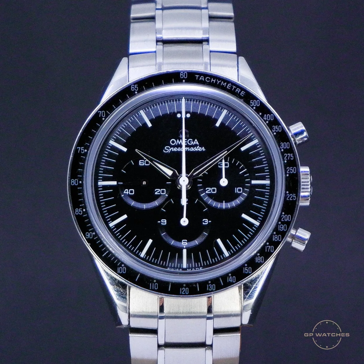 A close-up of a stainless steel Omega Speedmaster wristwatch with a black dial, three sub-dials, and a tachymeter scale on the bezel. The watch has a chronograph function and a silver bracelet.