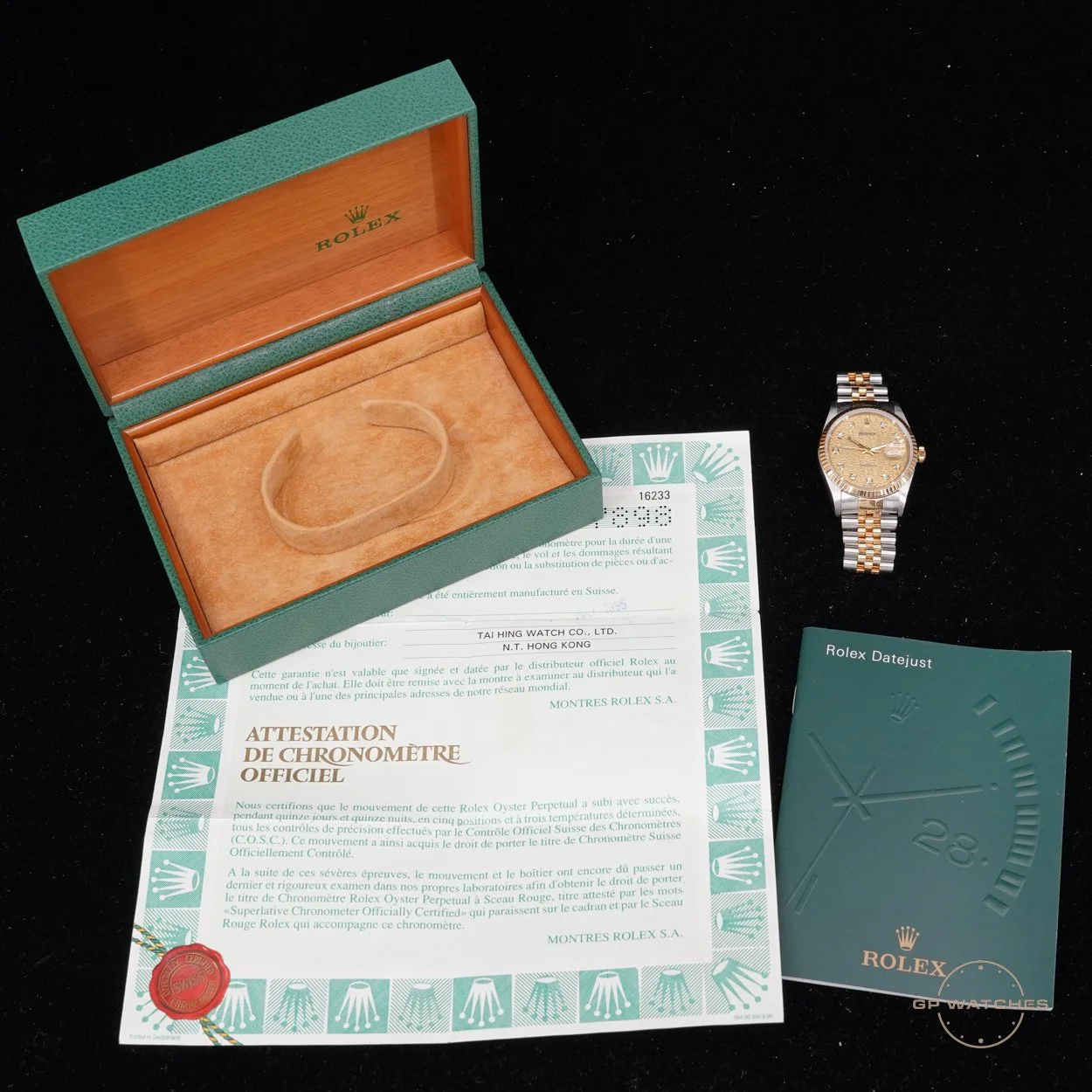 A Rolex watch with a two-tone metal band, a gold face, and a date display, placed next to its green Rolex box, certificate of authenticity, instruction booklet, and a transparent plastic watch holder on a black surface.
